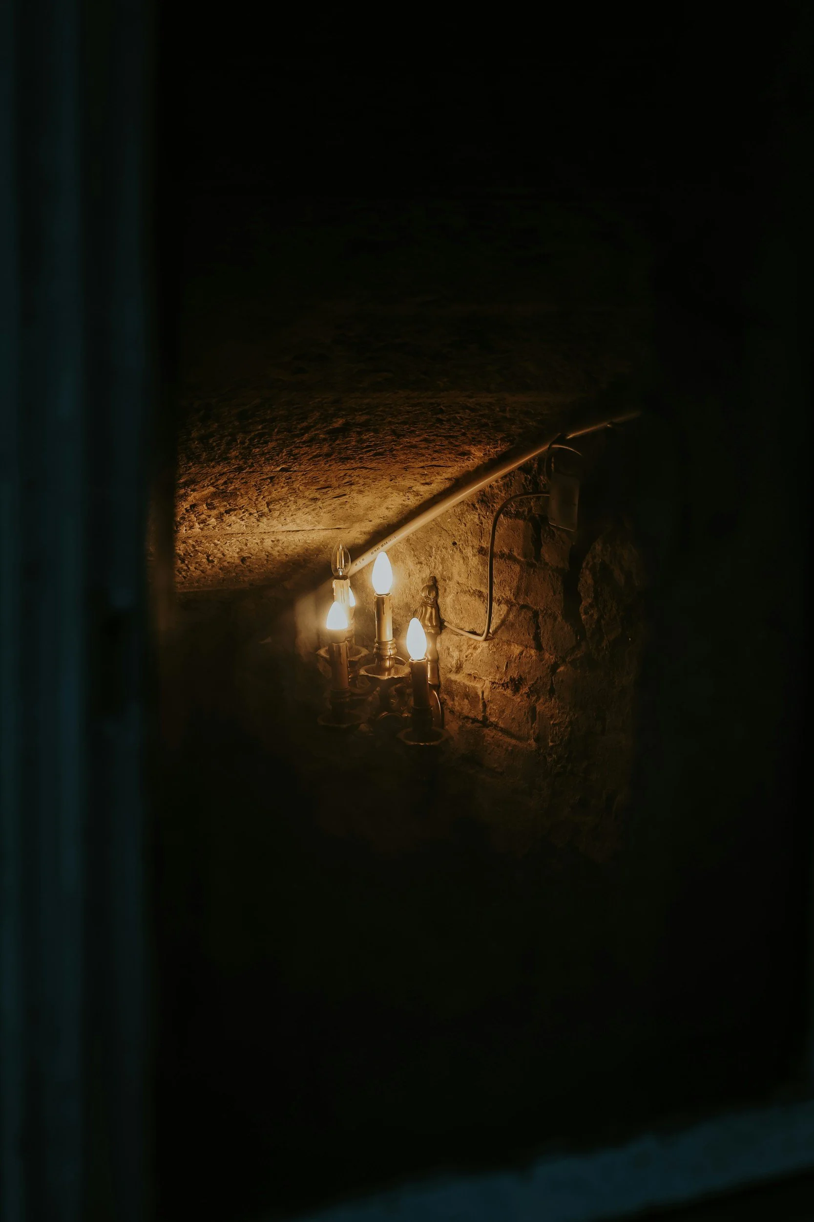An ornate metal candelabra holding lit candles attached to a dark, textured brick wall inside a dimly lit cellar