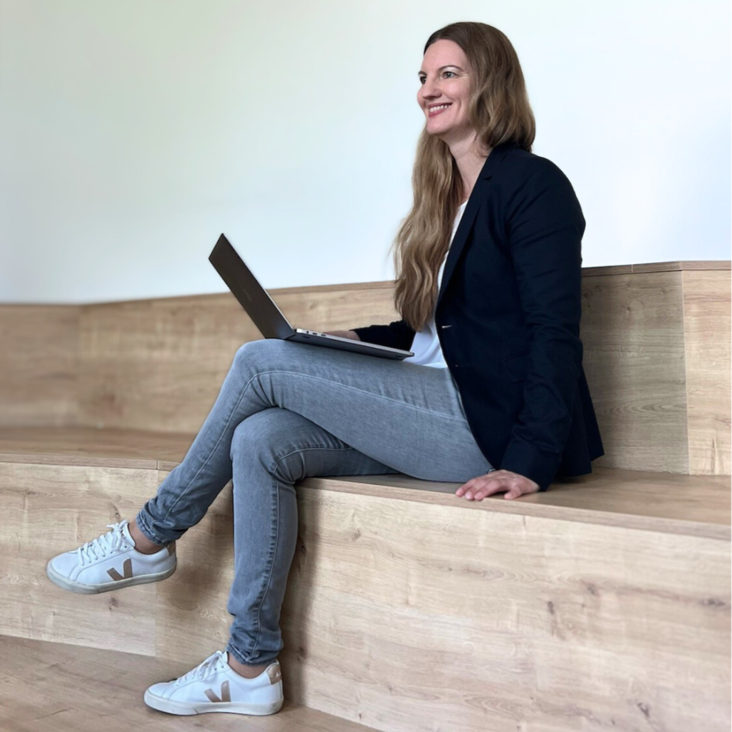 Frau sitzt auf einer Holzbank, benutzt einen Laptop, trägt eine dunkle Jacke, weiße Schuhe und hat lange braune Haare, lächelt und schaut nach rechts.