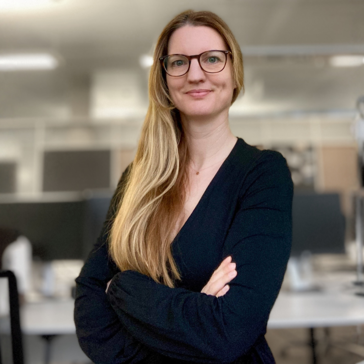 Eine Frau mit Brille und langem, glattem, blondem Haar, die lächelnd in einem Büro steht.