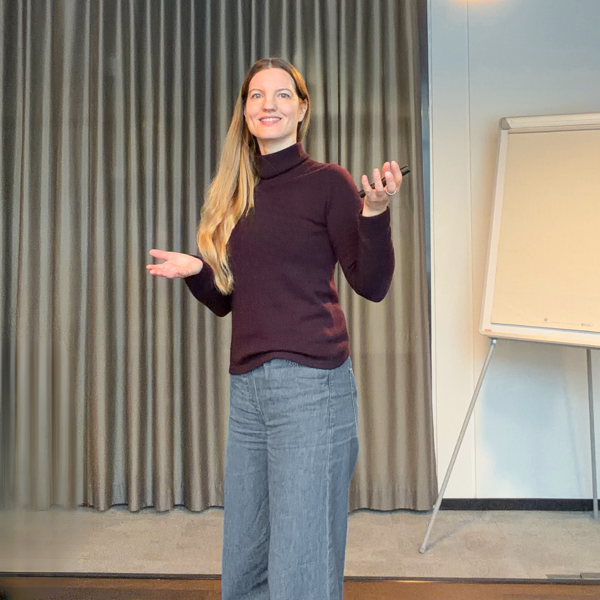 Eine Frau bei einer Präsentation hält ein Mikrofon in einer Hand, vor einem Flipchart, mit einem Vorhang im Hintergrund.
