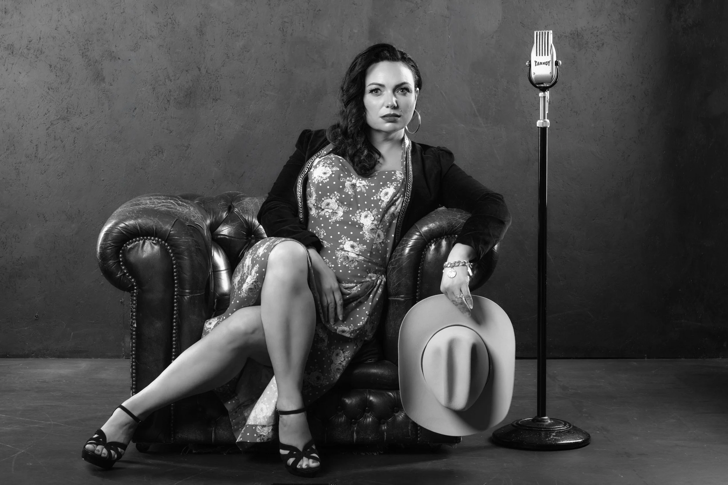 Black and white photograph of a woman sitting on a vintage leather couch, holding a wide-brimmed hat, with a microphone on a stand beside her, against a textured wall background.