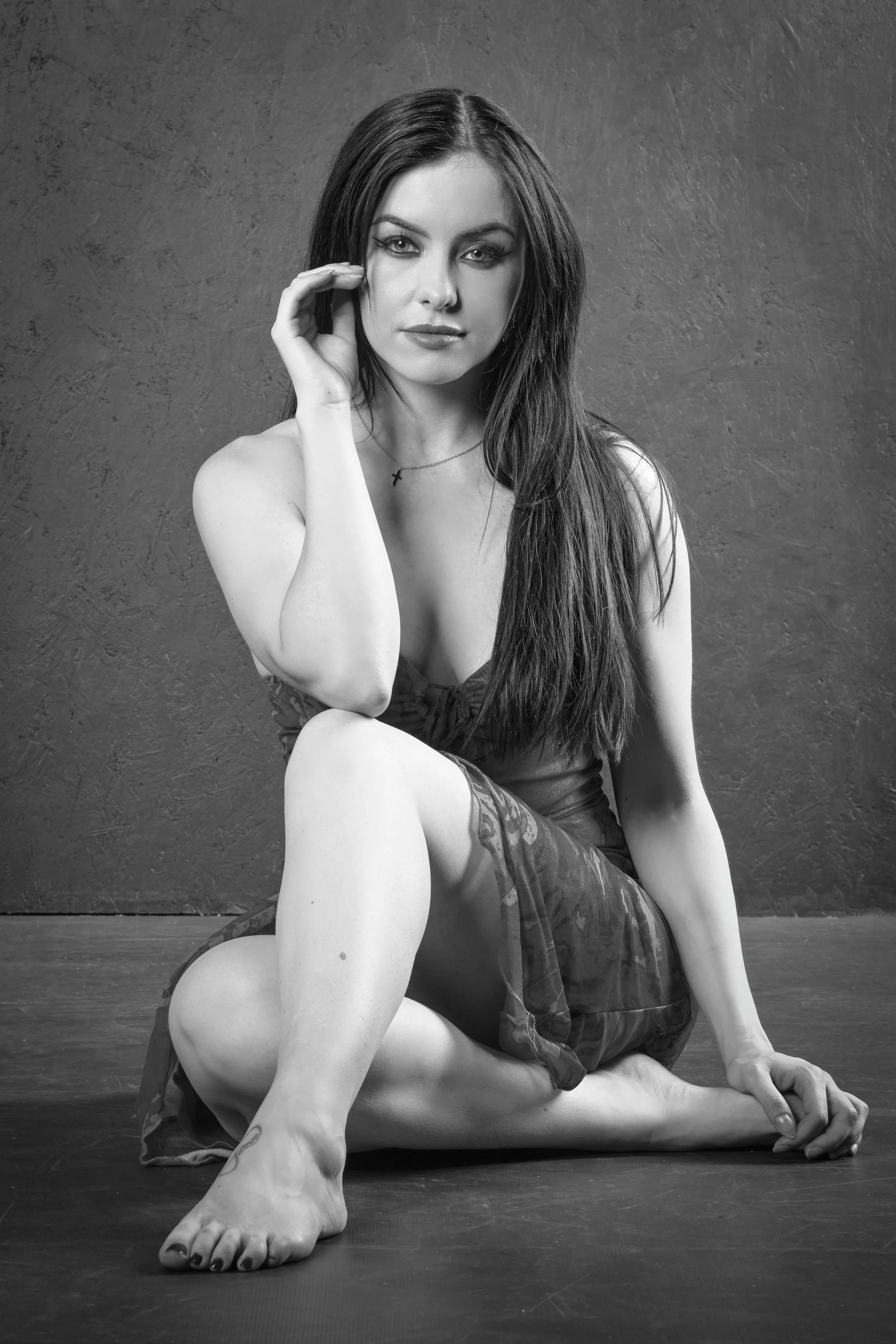Black and white photo of a woman with long dark hair, sitting on the floor with one leg bent and the other extended, touching her face with her hand, wearing a necklace with a cross. She is dressed in a sleeveless top and a short skirt, against a textured wall background.