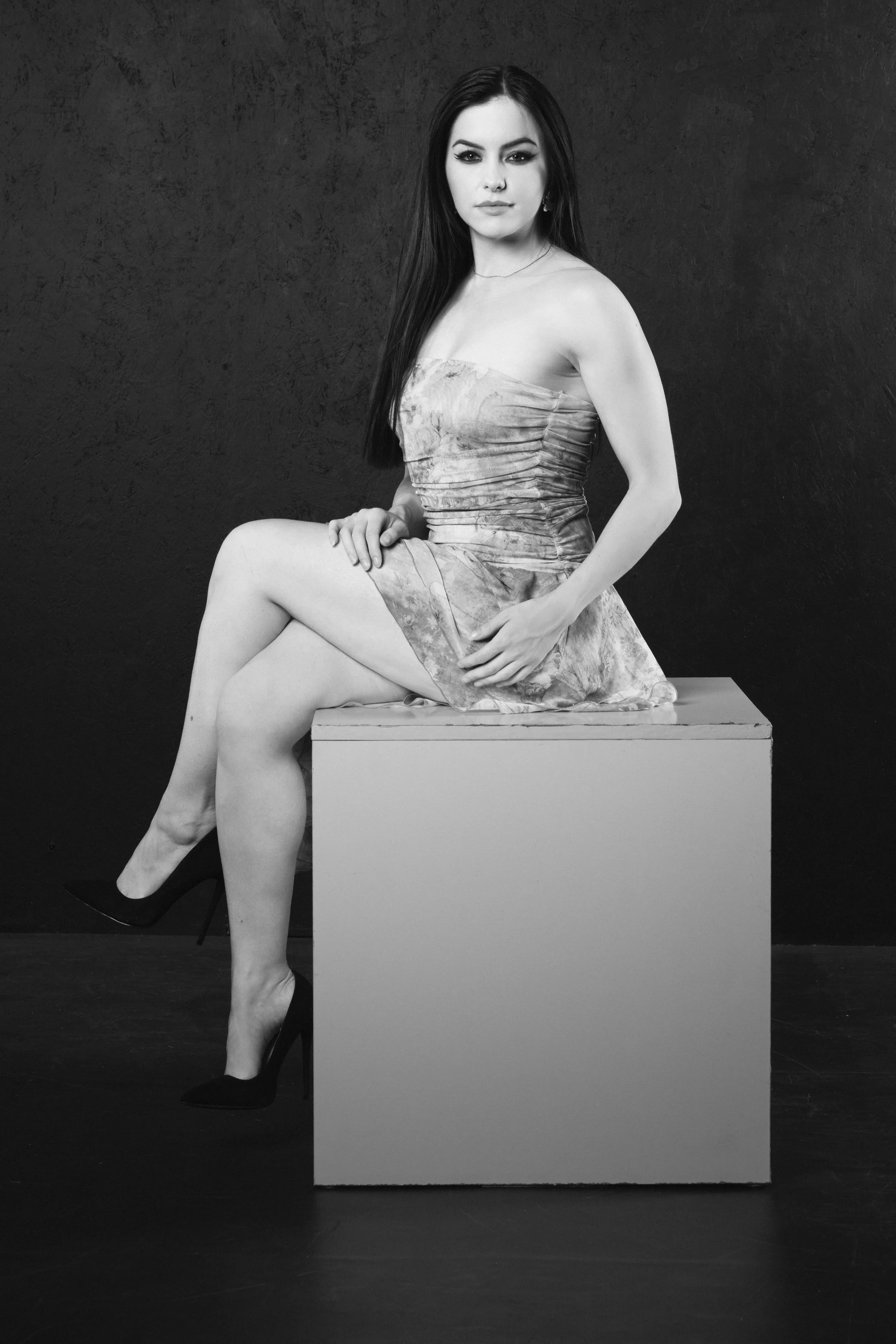 A black-and-white photograph of a woman sitting on a box, wearing a strapless dress and high heels, with a serious expression on her face and dark hair over her shoulder, against a textured dark background.