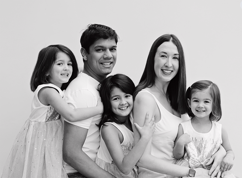 Family of five, includes two adult women and three children, smiling and sitting close together against a plain background.