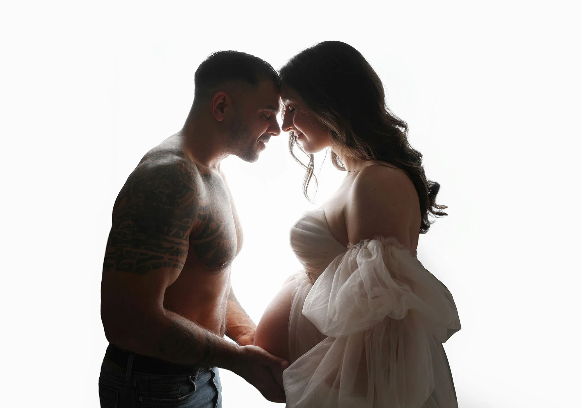 A couple touching foreheads, smiling, with a pregnant woman wearing a light dress and a man with tattoos on his arm, against a white background.