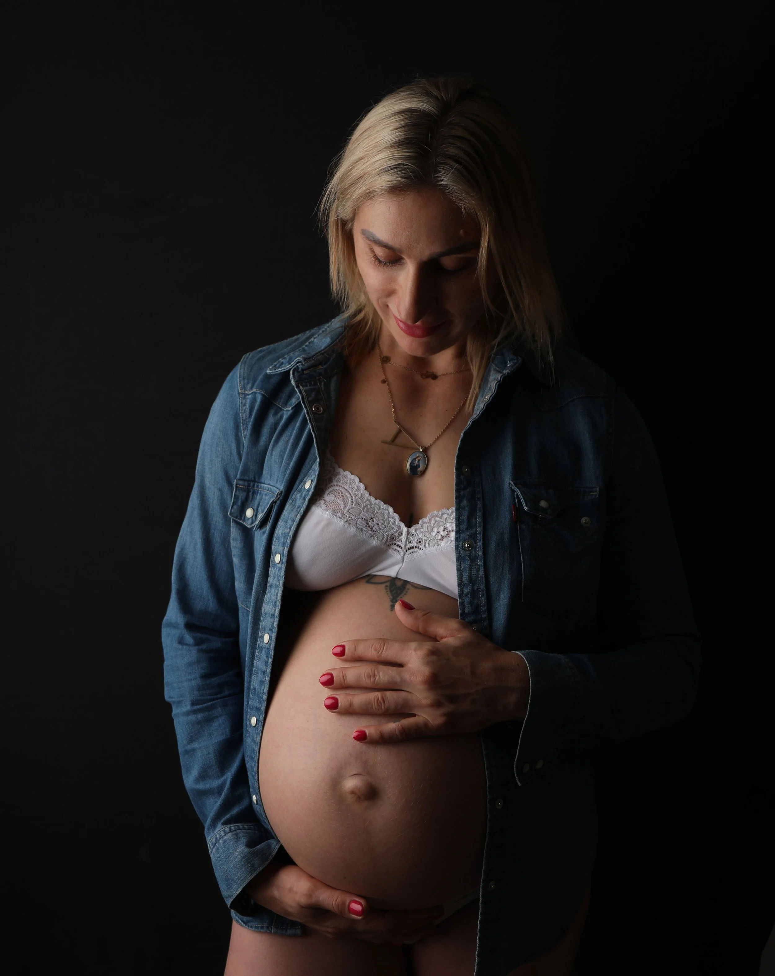 A pregnant woman with blonde hair wearing a denim jacket and white lace top, gently holding her exposed belly, with a dark background.