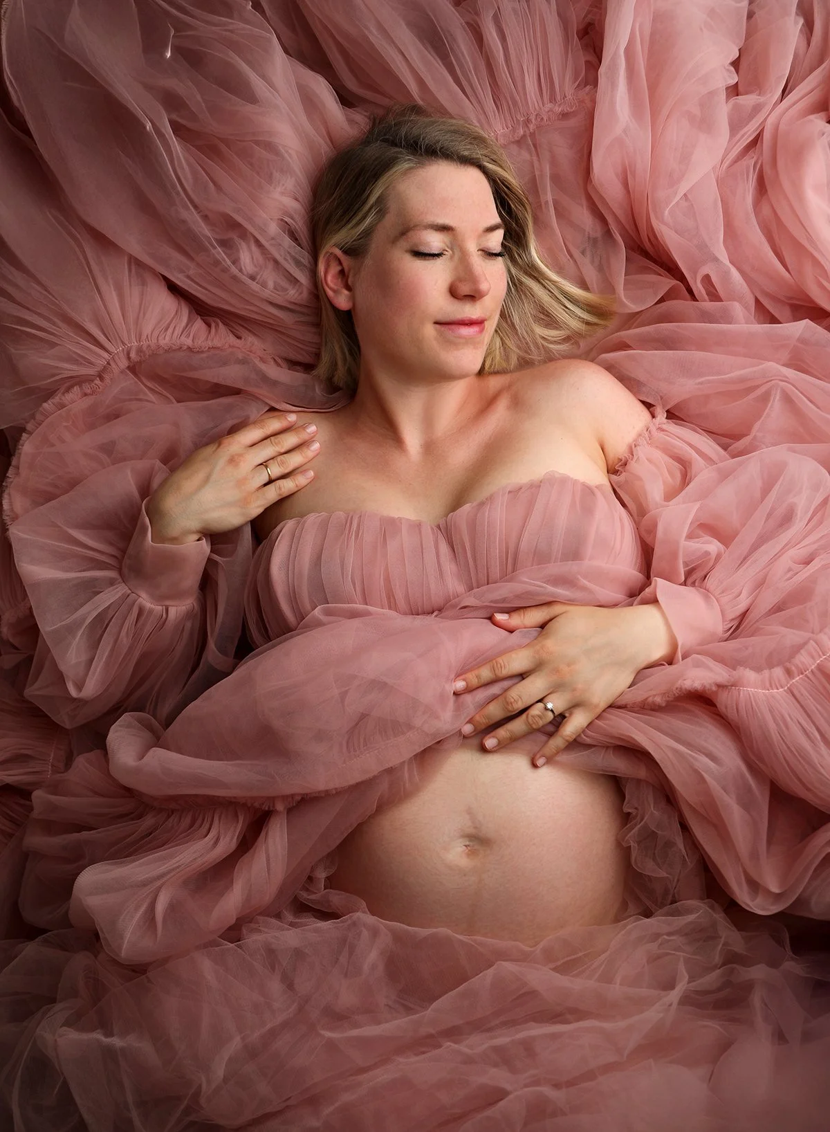 A pregnant woman with blonde hair, lying on a bed of pink tulle fabric. She is wearing a pink, off-the-shoulder dress with a ruched bodice. Her eyes are closed, and she has a peaceful expression on her face. She is gently touching her baby bump with 