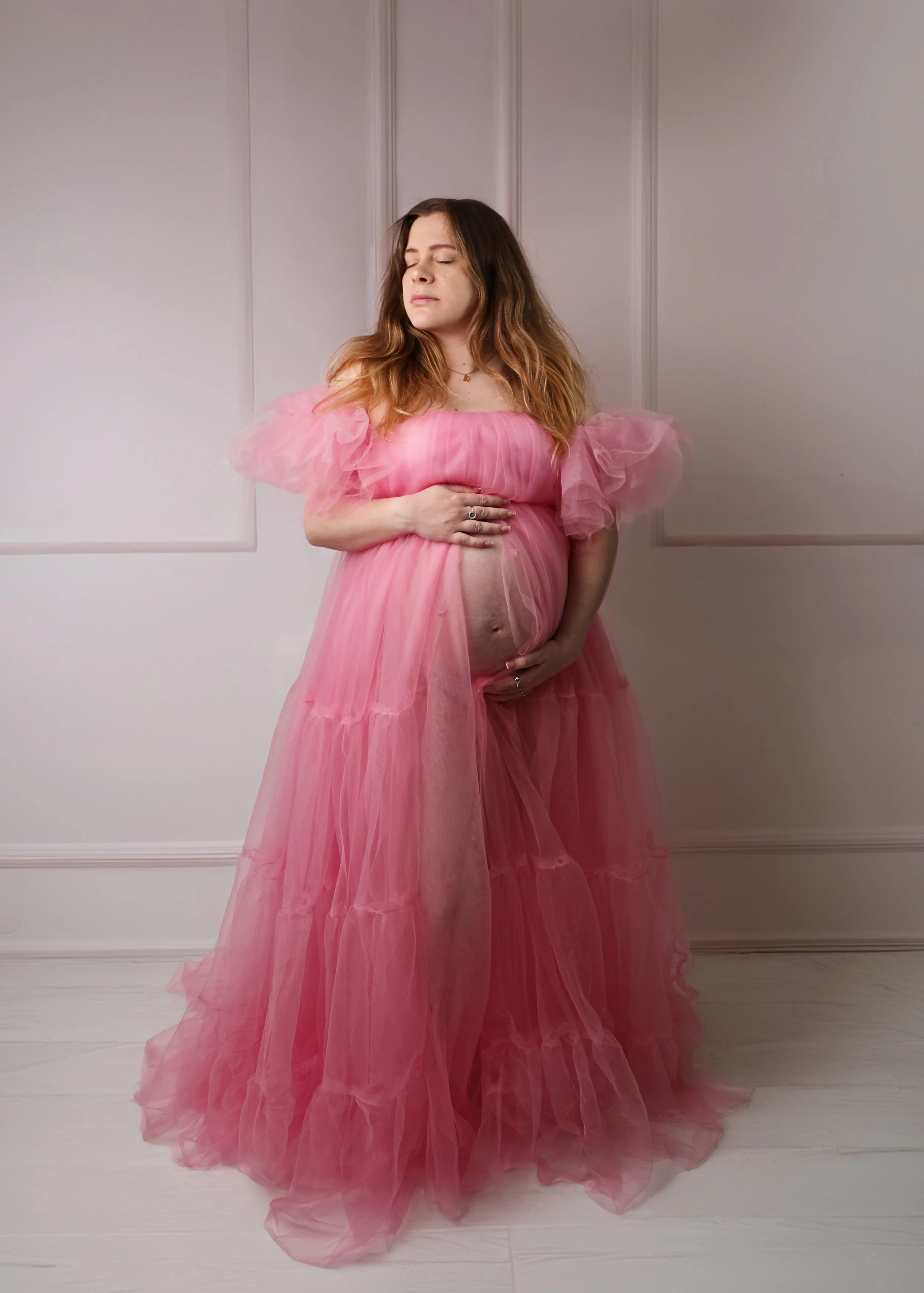 A woman with long, wavy brown hair, wearing a flowing pink dress with ruffled, sheer fabric sleeves, standing against a plain white wall with decorative paneling. She is holding her pregnant belly with one hand and has her eyes closed with a serene e