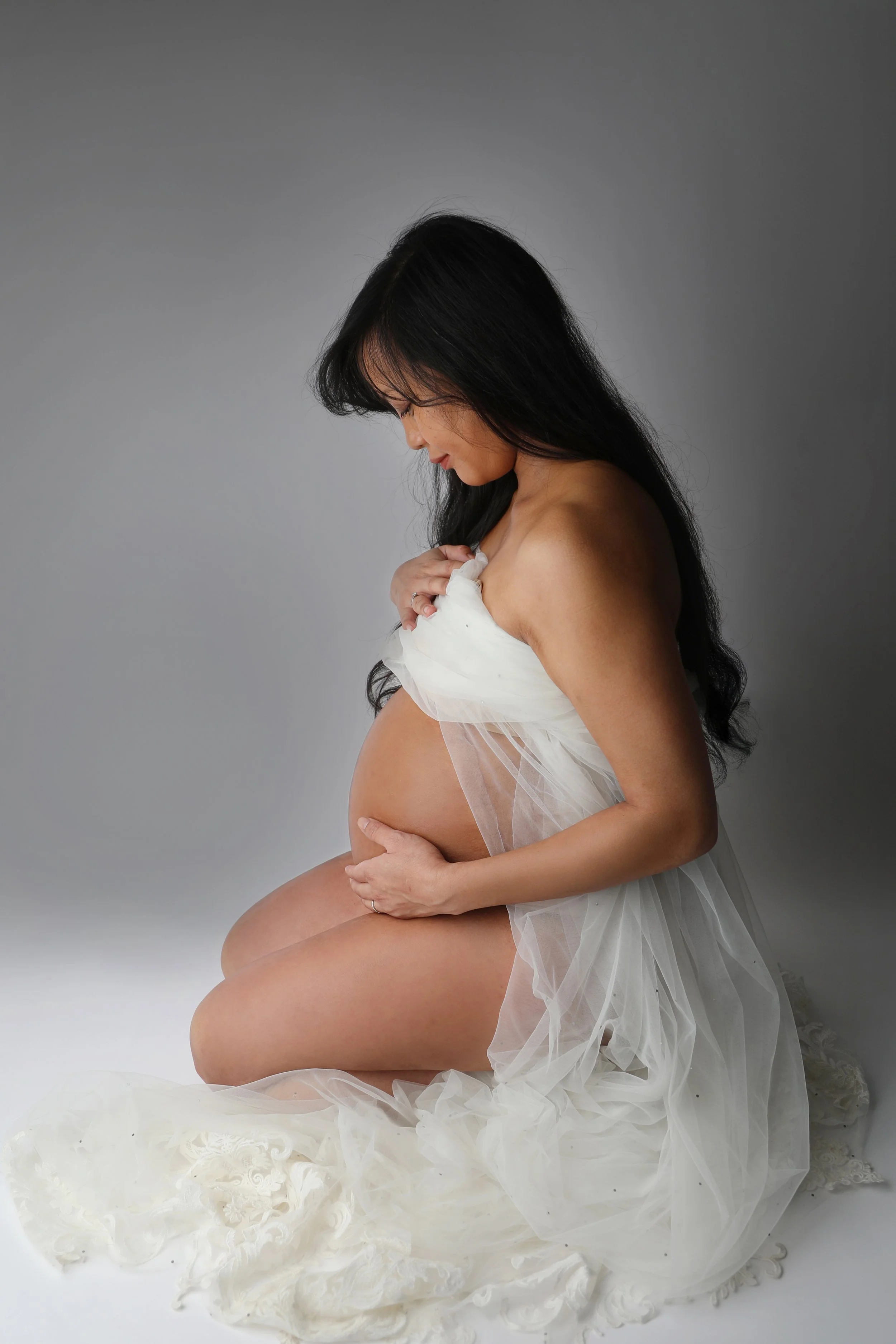 Pregnant woman with long black hair, wearing a sheer white tulle dress, sitting against a plain gray background, gently cradling her belly with one hand and holding a piece of fabric near her shoulder with the other.