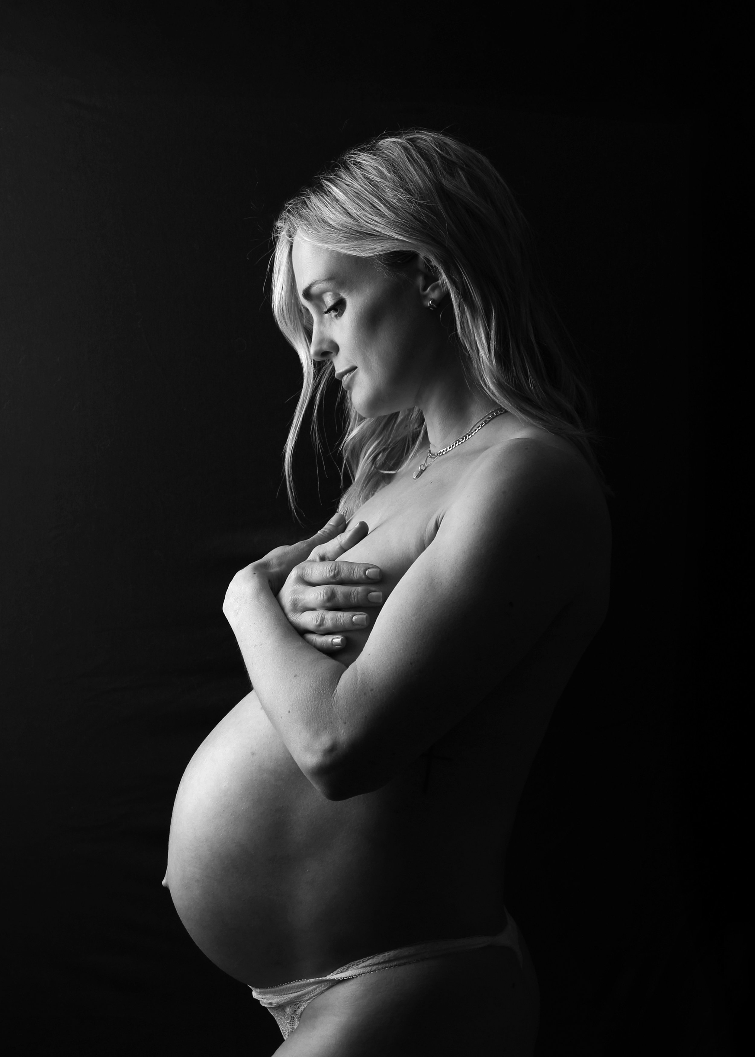 Black and white photo of a pregnant woman with her eyes closed, embracing her belly with both hands, wearing lace underwear, minimal jewelry, and standing against a dark background.