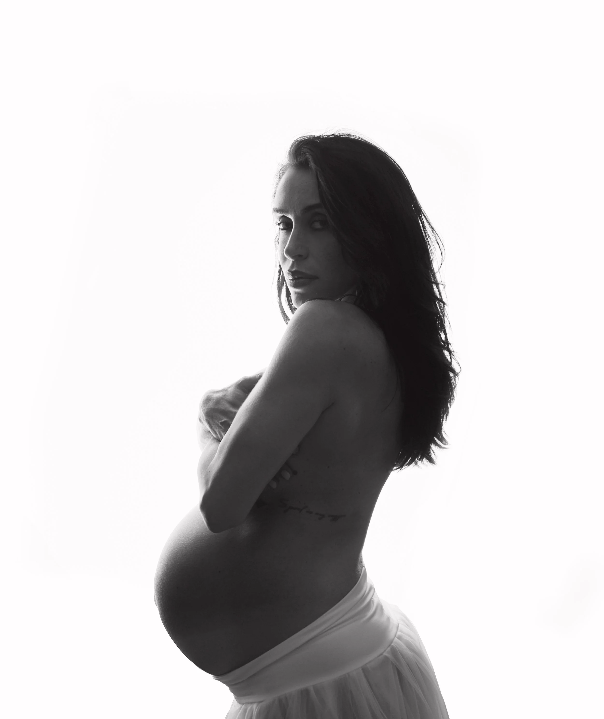 Black and white photo of a pregnant woman with long dark hair, standing sideways with arms crossed, wearing a light skirt and no top, against a plain white background.