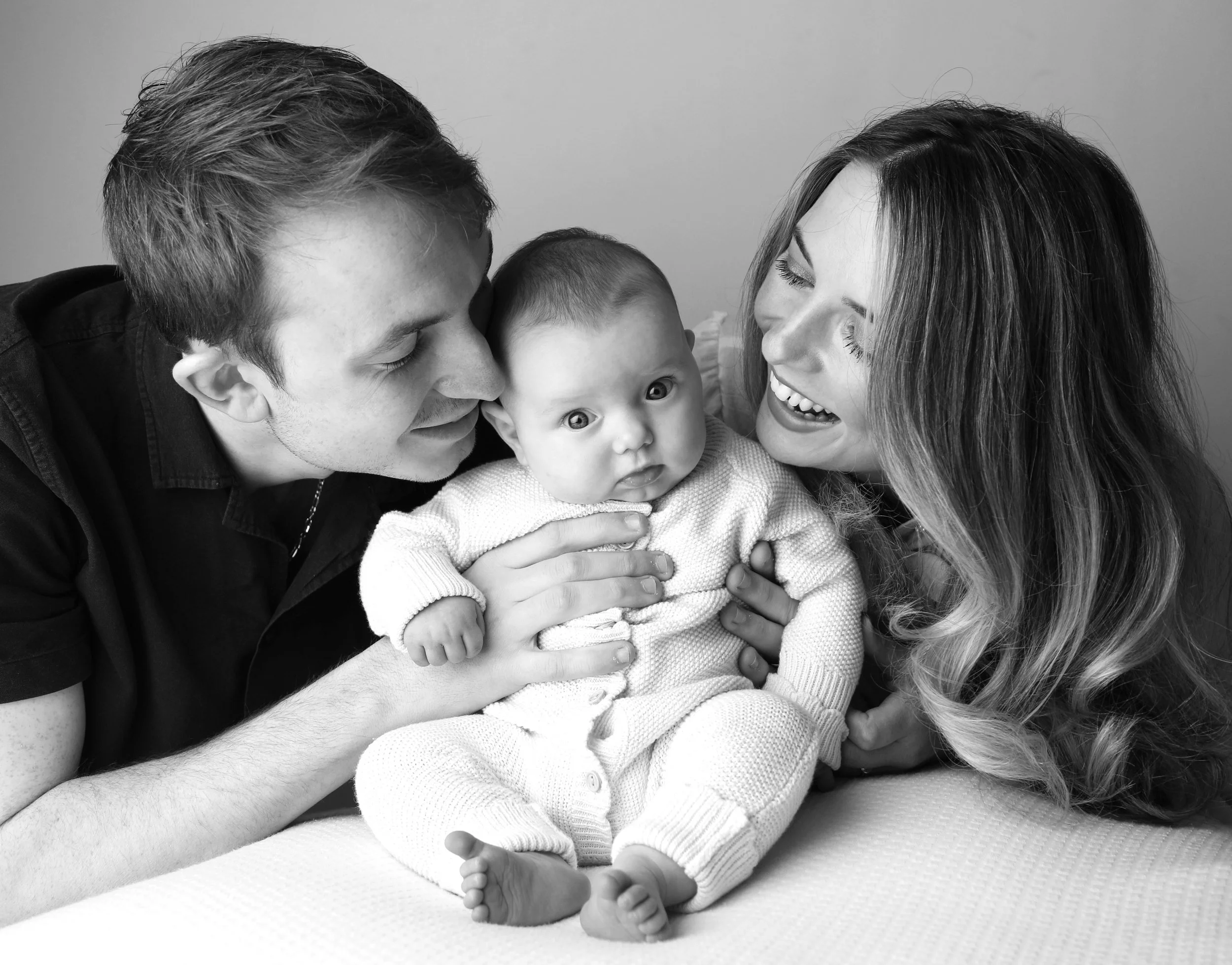A black and white photo of a family of three, with a man and woman happily holding and leaning towards their baby, who is sitting on a surface and looking at the camera.