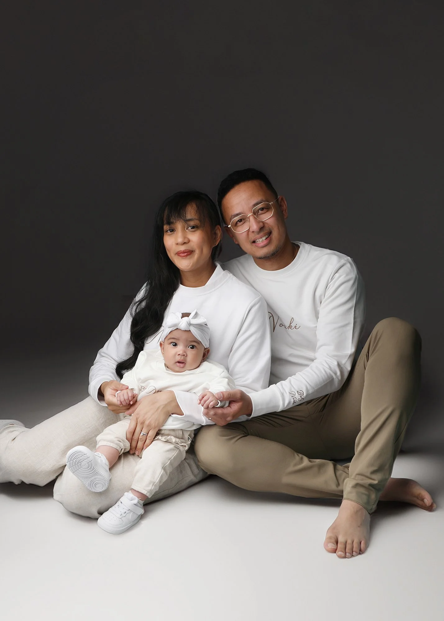 A family of four posing together against a dark background. The woman and man are sitting, with the woman holding a baby on her lap. The man is smiling, wearing glasses, and has his arm around the woman. The woman has long, dark hair, and the baby is wearing a headband with a bow. The family appears relaxed and happy.