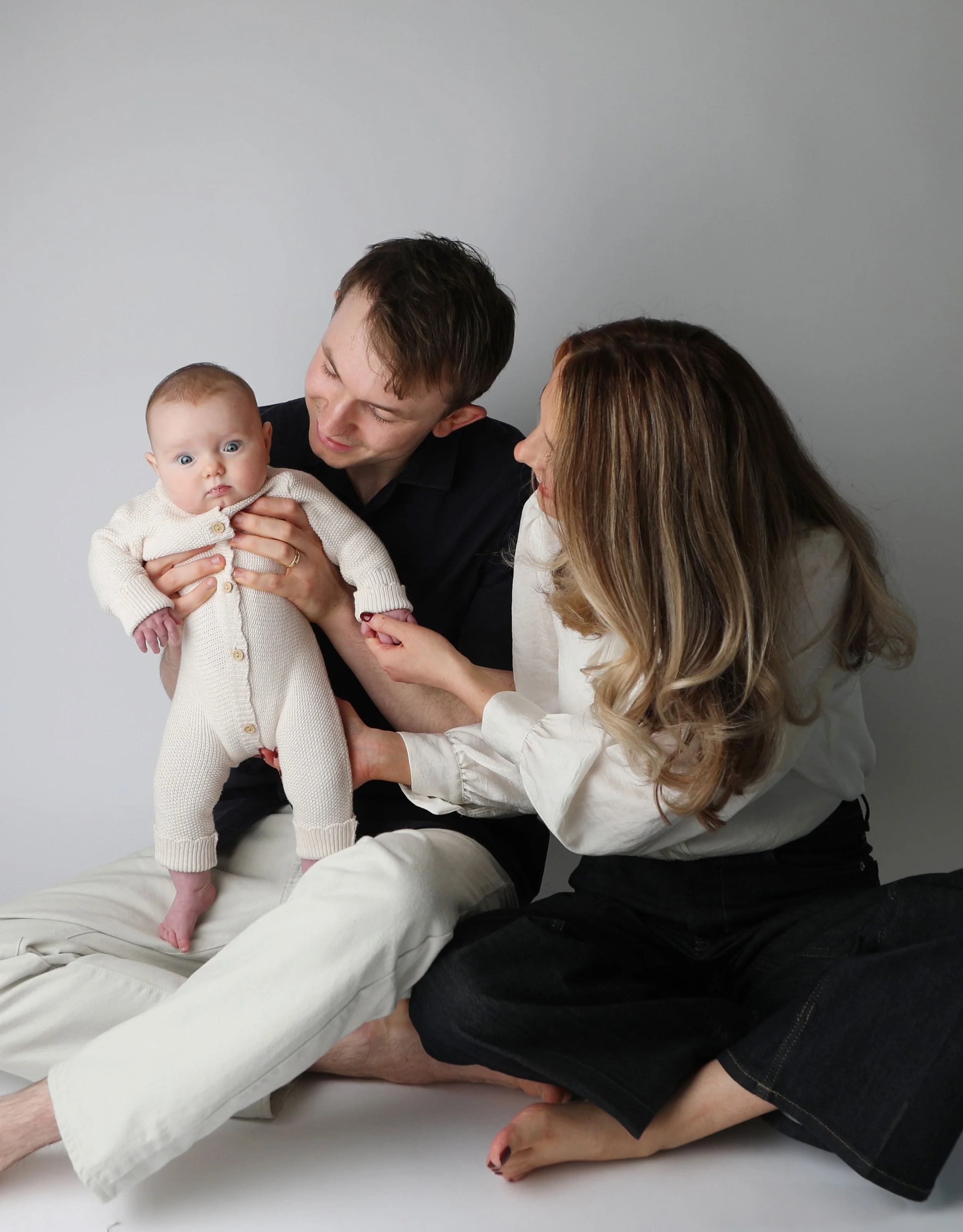 Family of three, a man, woman, and a baby, interacting against a plain light-colored background.
