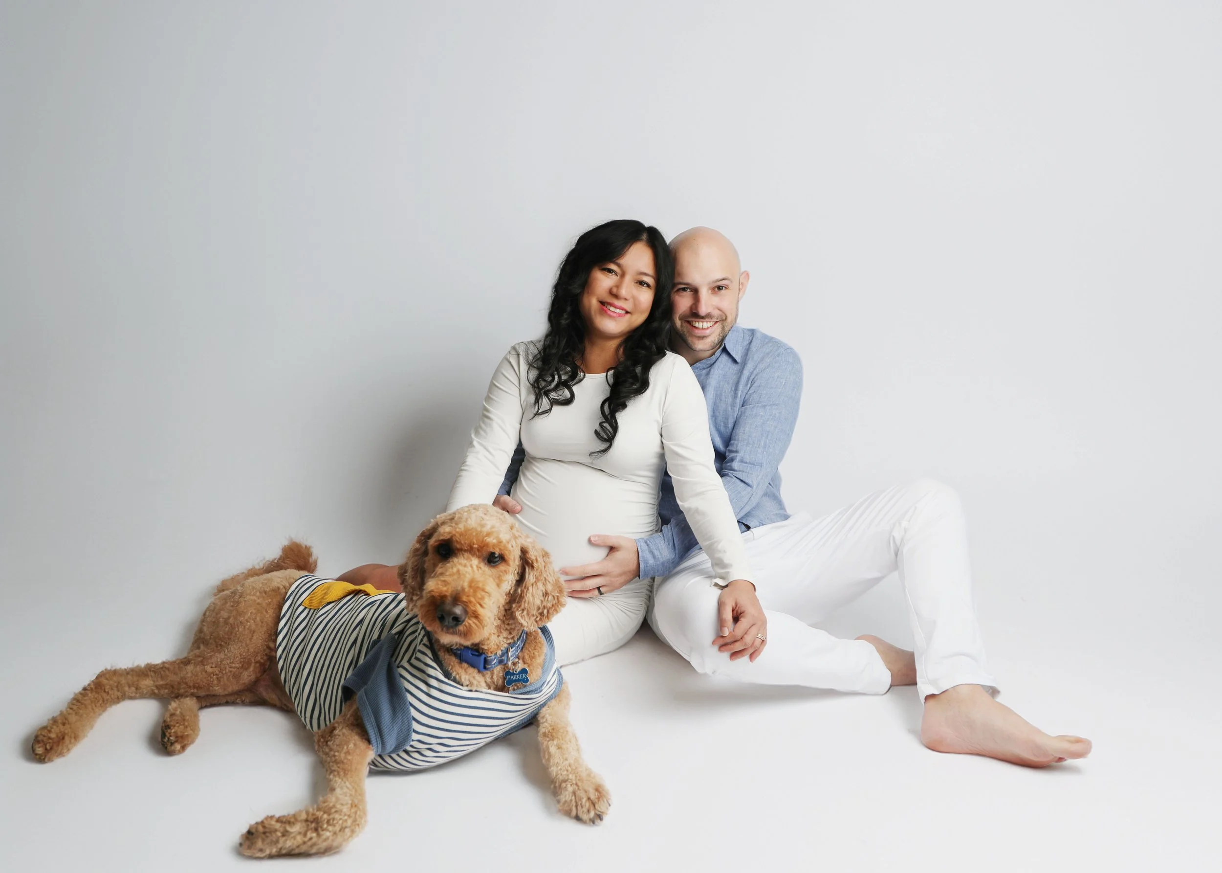 A couple sitting on the floor with their dog, pregnant woman smiling, man hugging her, dog wearing striped shirt and blue collar, another dog lying down, plain light gray background.