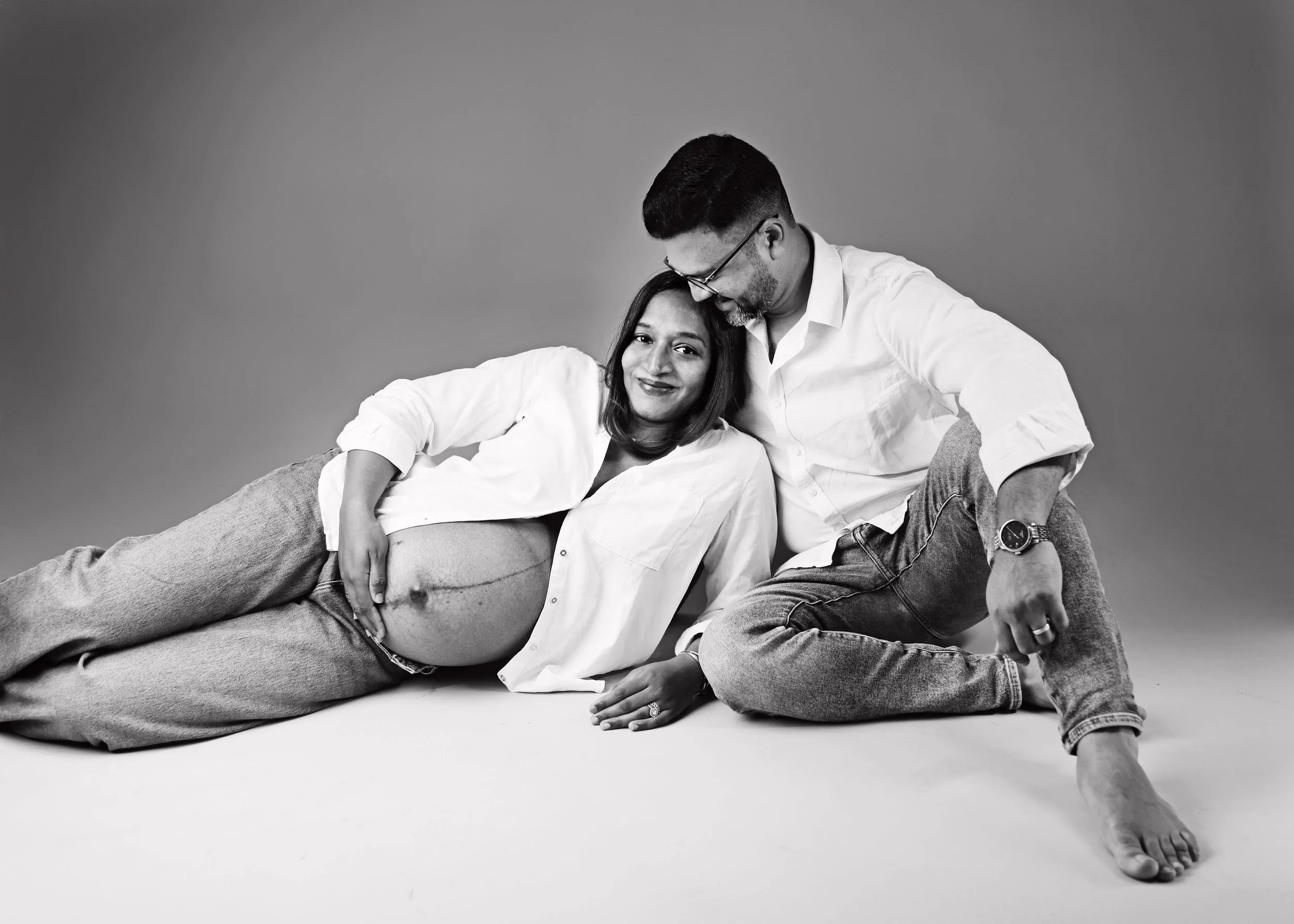 Black and white photo of a smiling woman lying on the ground with her head on a man's lap, and a man leaning over her, both dressed casually, against a plain background.