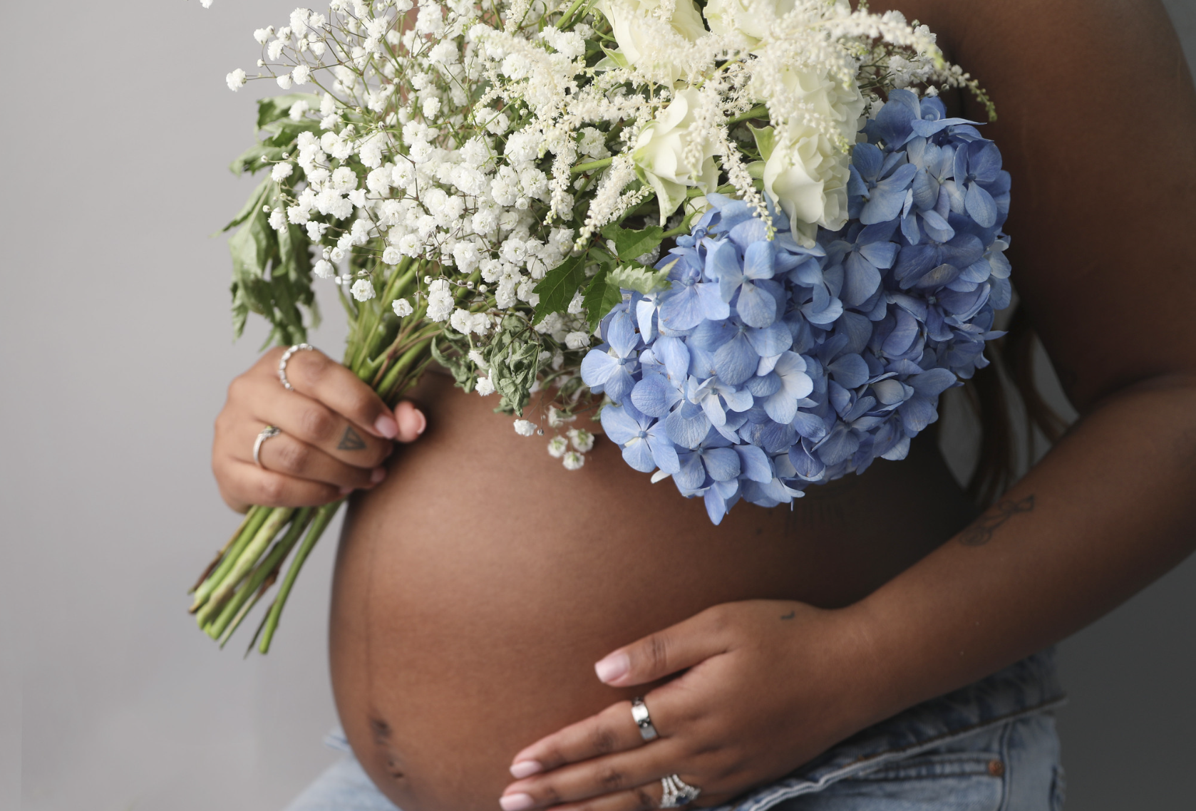A pregnant woman holding a bouquet of white and blue flowers, with visible tattoos and rings on her fingers, against a plain gray background.
