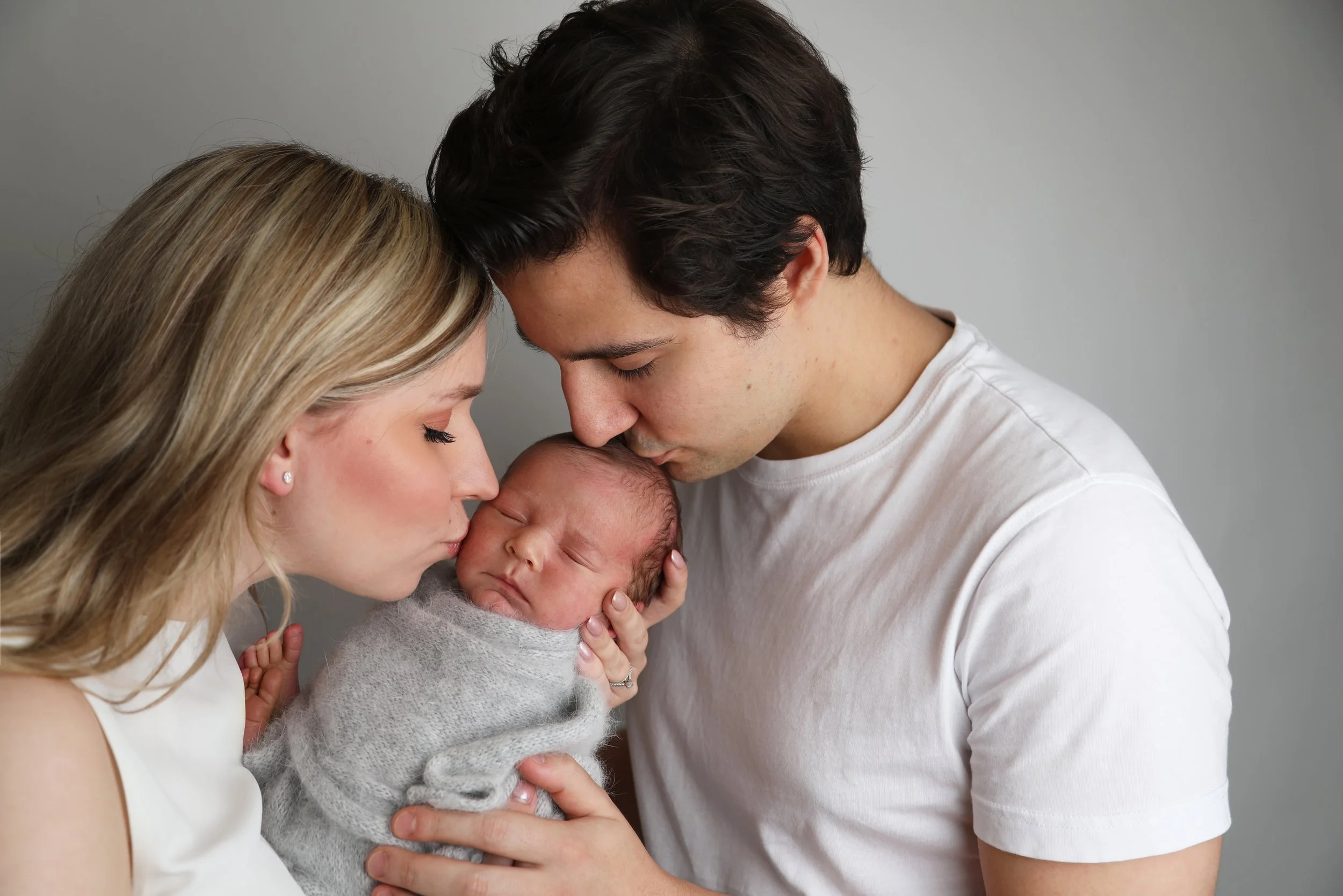 A woman and a man gently kissing a sleeping newborn wrapped in a gray blanket.