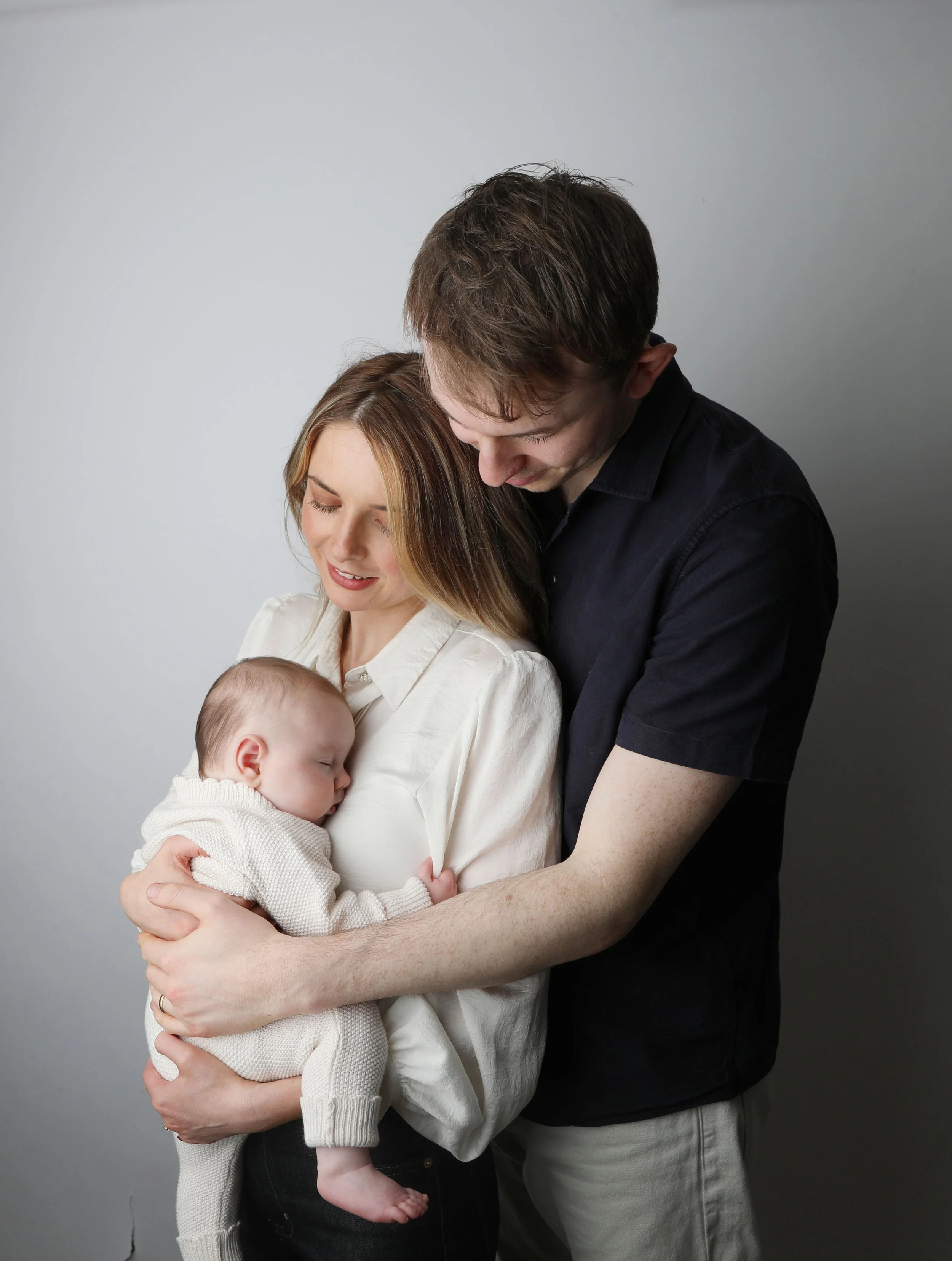 A young couple holding their sleeping baby, the woman is smiling, and the man is looking at the baby, all against a plain light gray background.