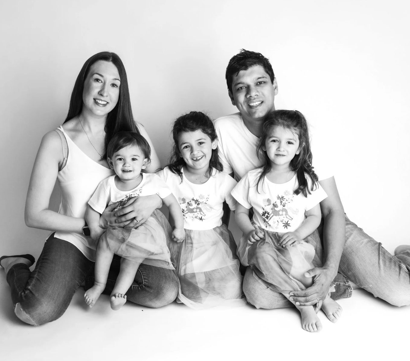 A black and white photo of two adults and three young girls sitting together on the floor, smiling at the camera.