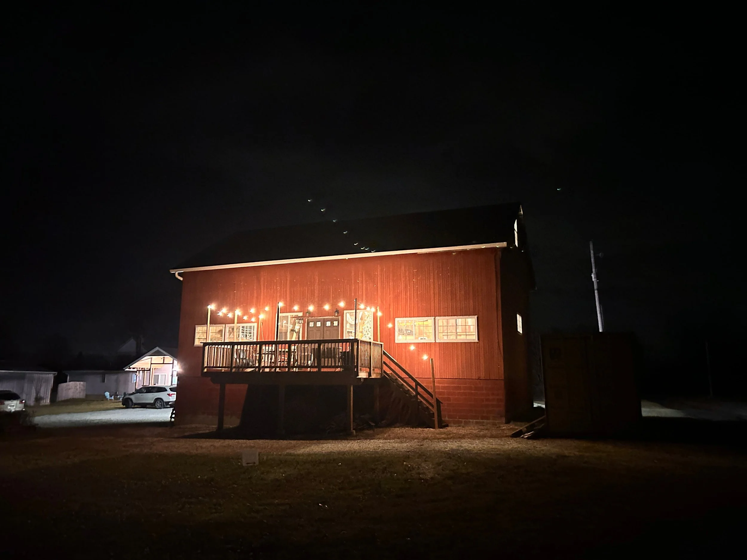 A red barn illuminated with string lights at night, with a small deck and stairs leading up to the entrance.