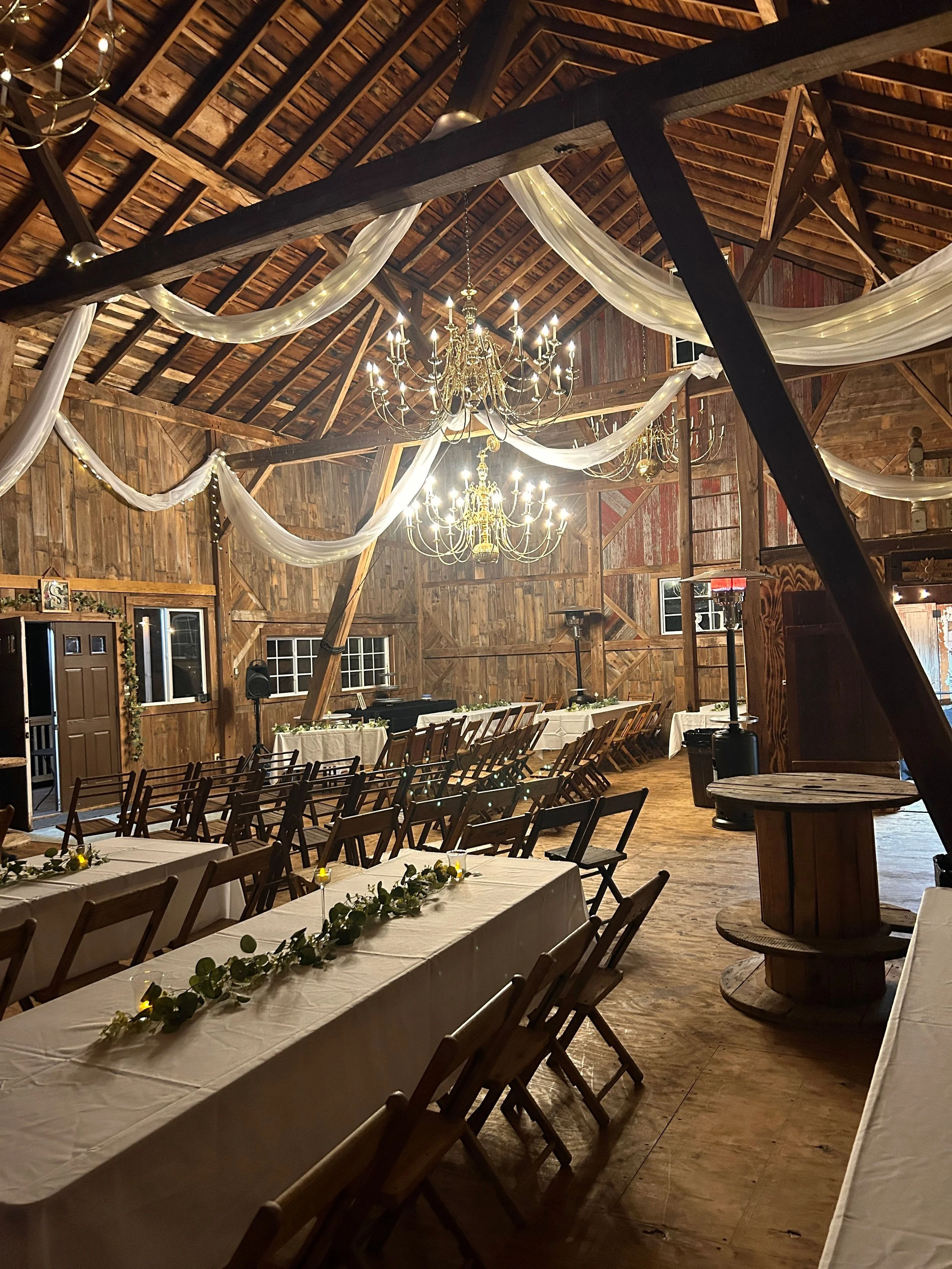 Rustic barn interior decorated for an event with chandeliers, white drapery, and long tables with white tablecloths and floral centerpieces.