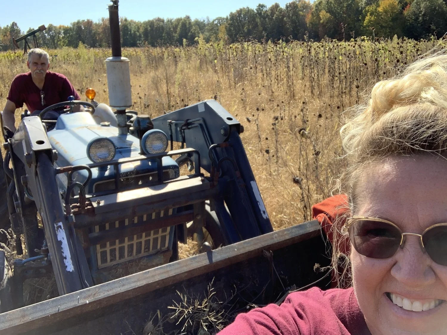 A woman with blonde curly hair wearing sunglasses and a maroon shirt takes a selfie in a field of dry crops, with a man on a tractor behind her in a red shirt, set in a sunny rural area with trees in the background.
