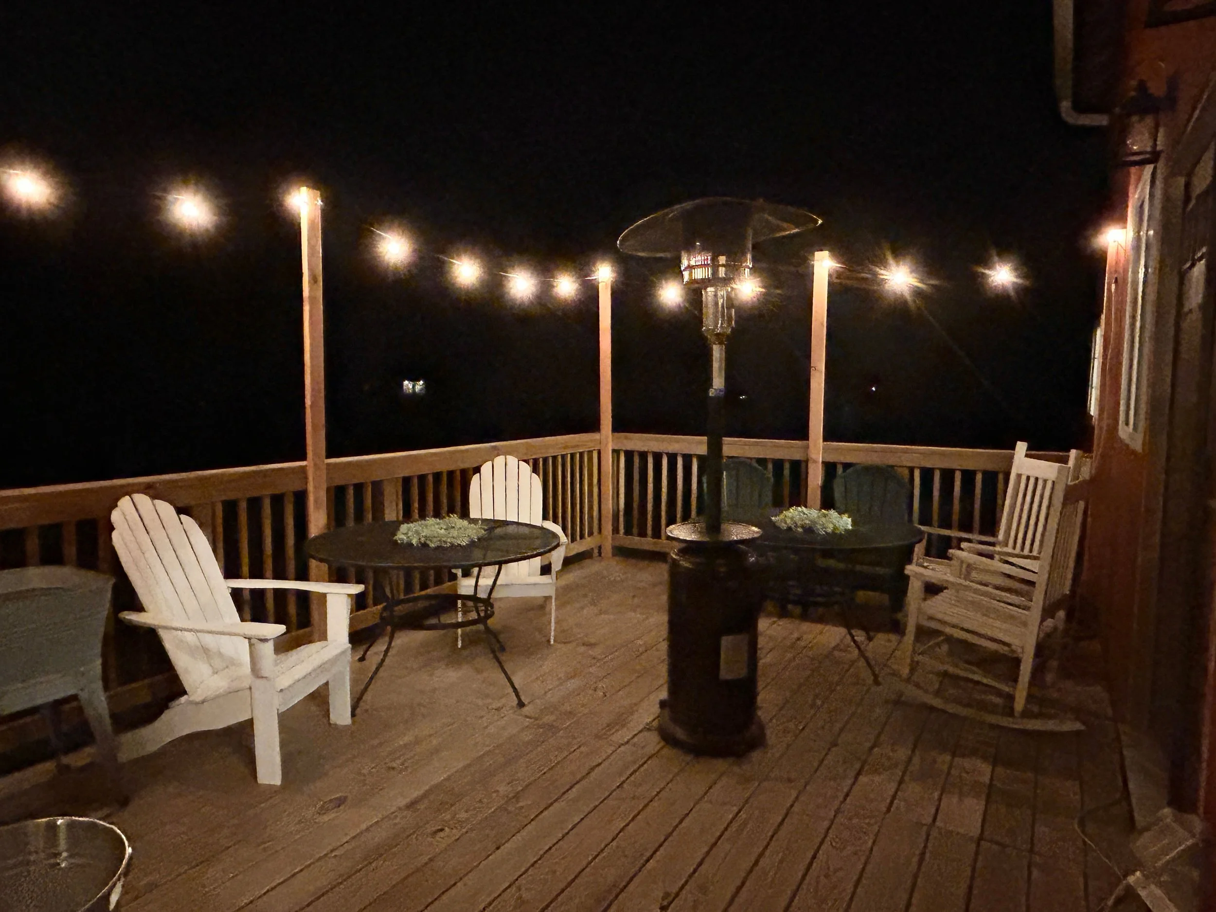 A wooden deck at night with string lights overhead, outdoor chairs, a small round table with potted plants, a patio heater, and a wooden fence.