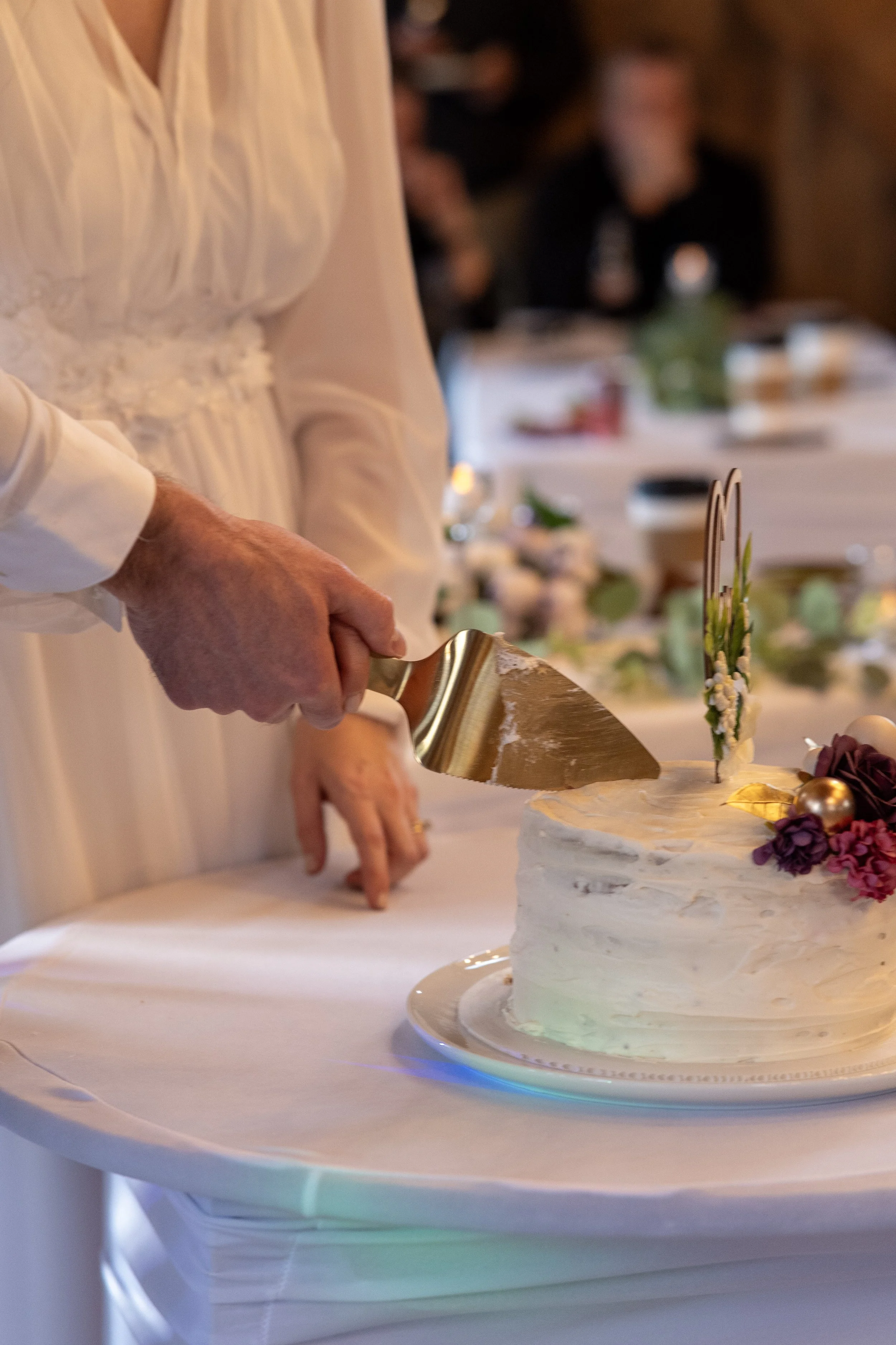 Person in a white dress cutting a wedding cake decorated with flowers and gold ornaments at a wedding reception.