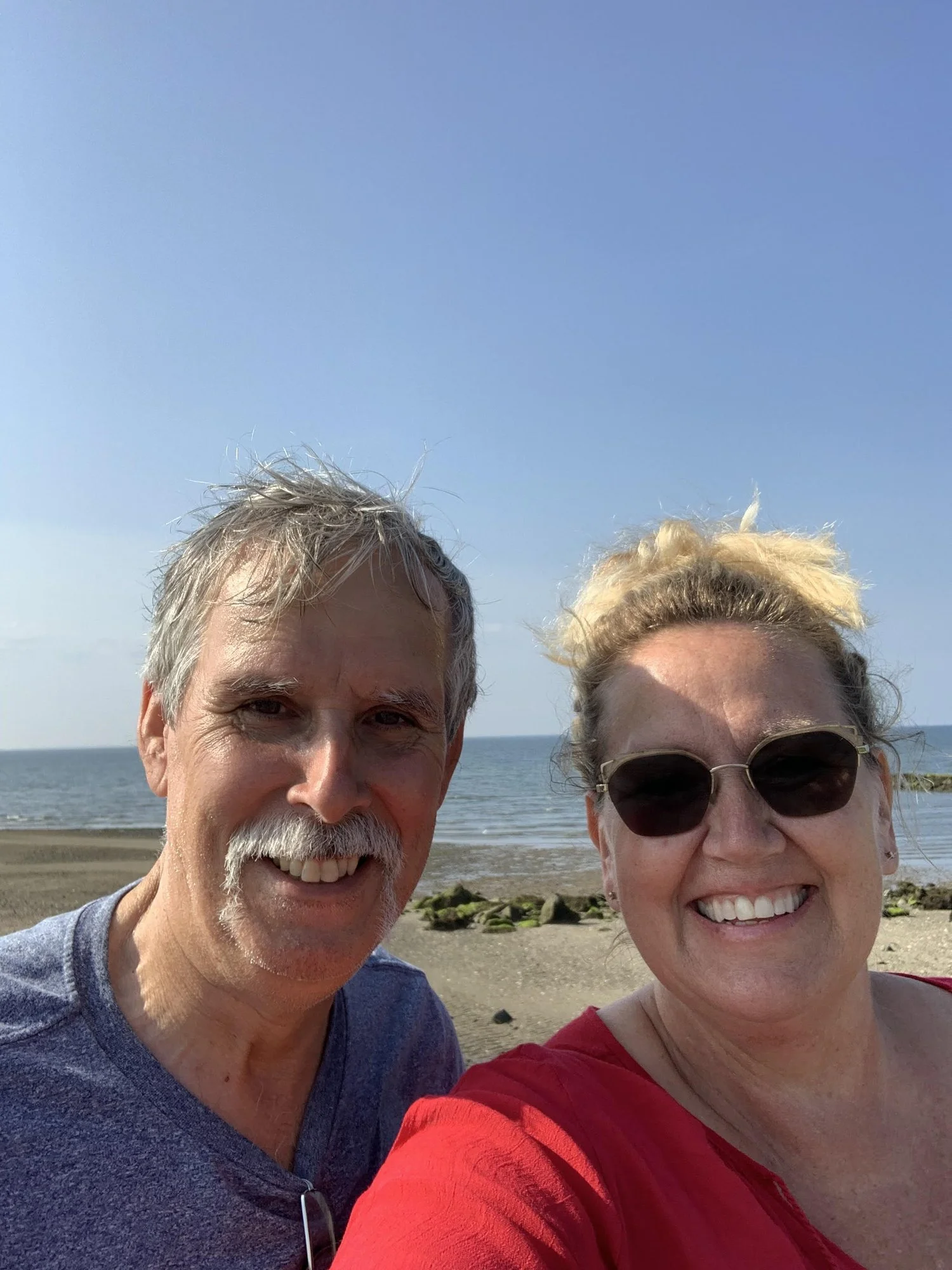 A smiling man and woman taking a selfie on a beach with the ocean and rocks in the background.