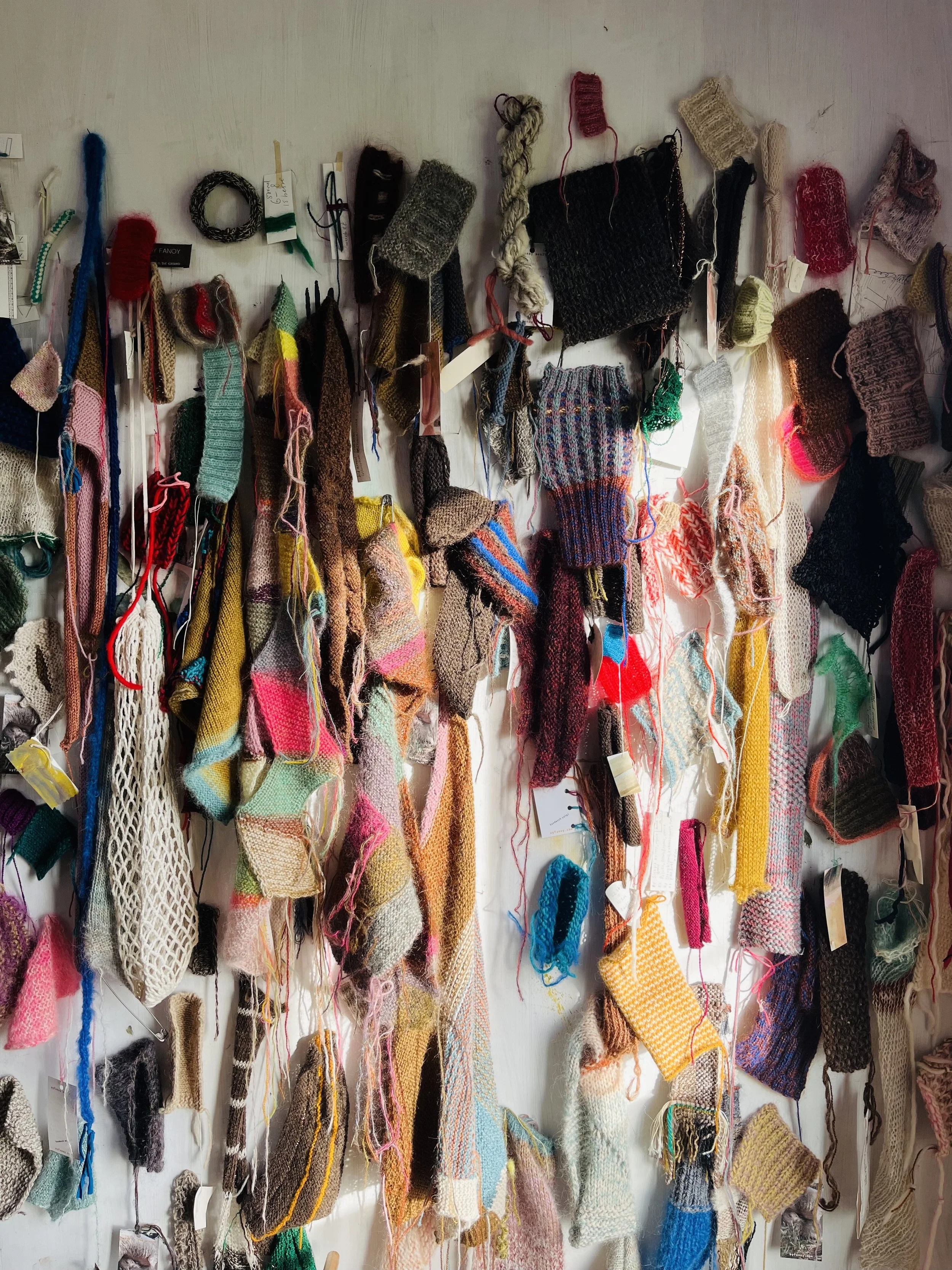 A wall covered with various colorful hand-knit and crocheted sock samples, some with tags, hanging and pinned in a display.