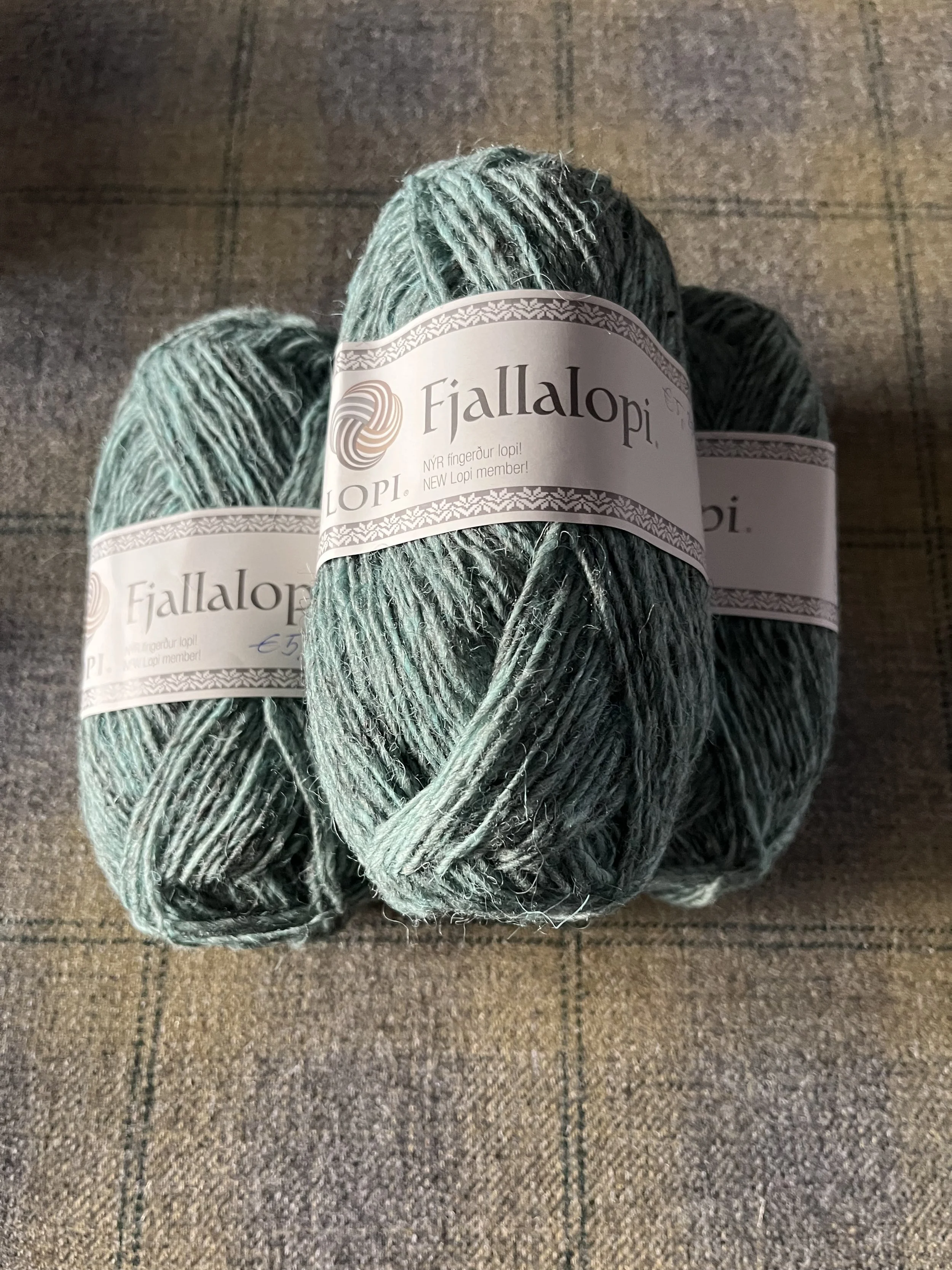 Three balls of Fjalllopi yarn in shades of green and gray on a wooden surface.