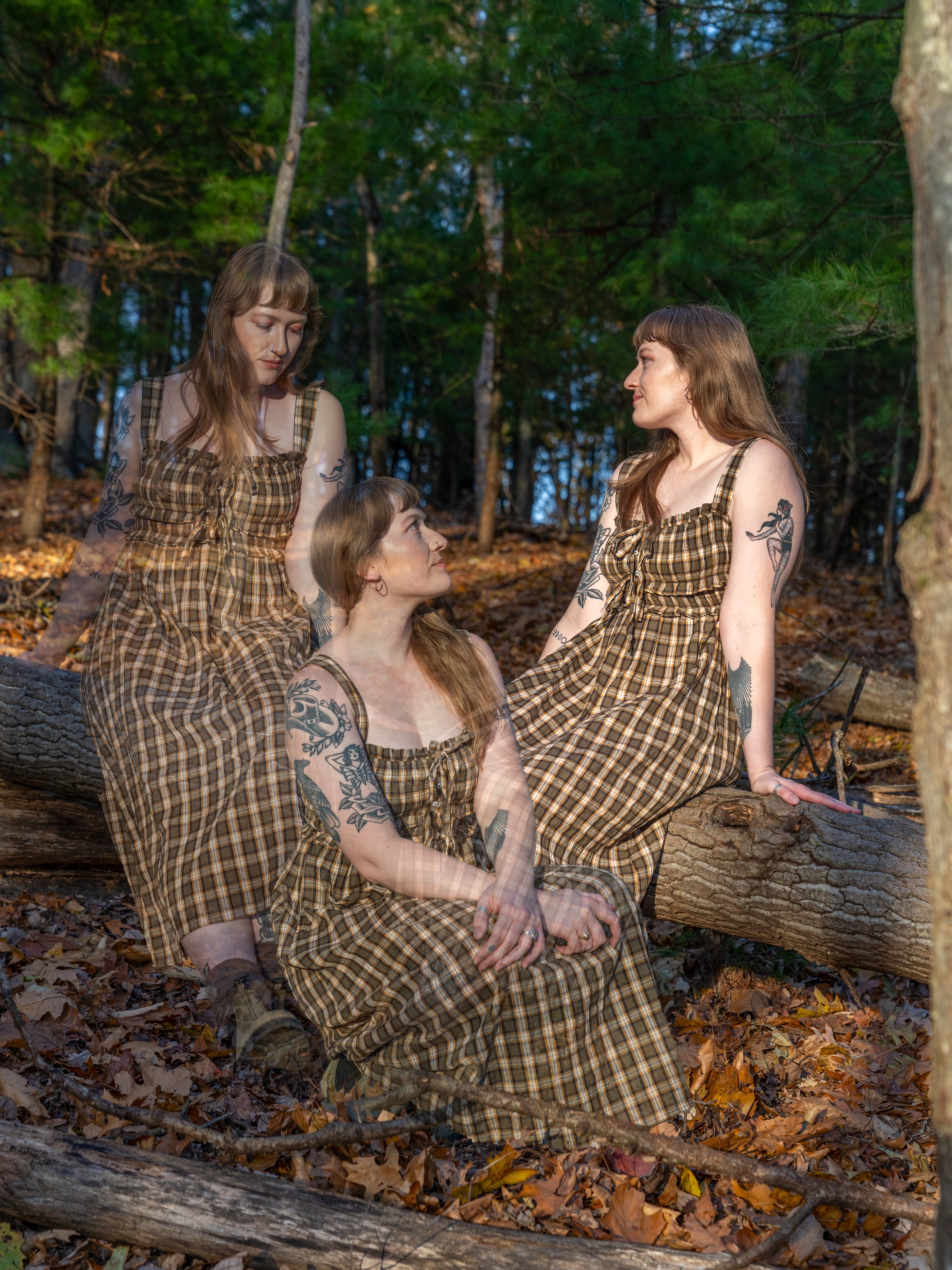 Four women with tattoos wearing matching plaid dresses sitting on a fallen tree in a forest with trees and autumn leaves.
