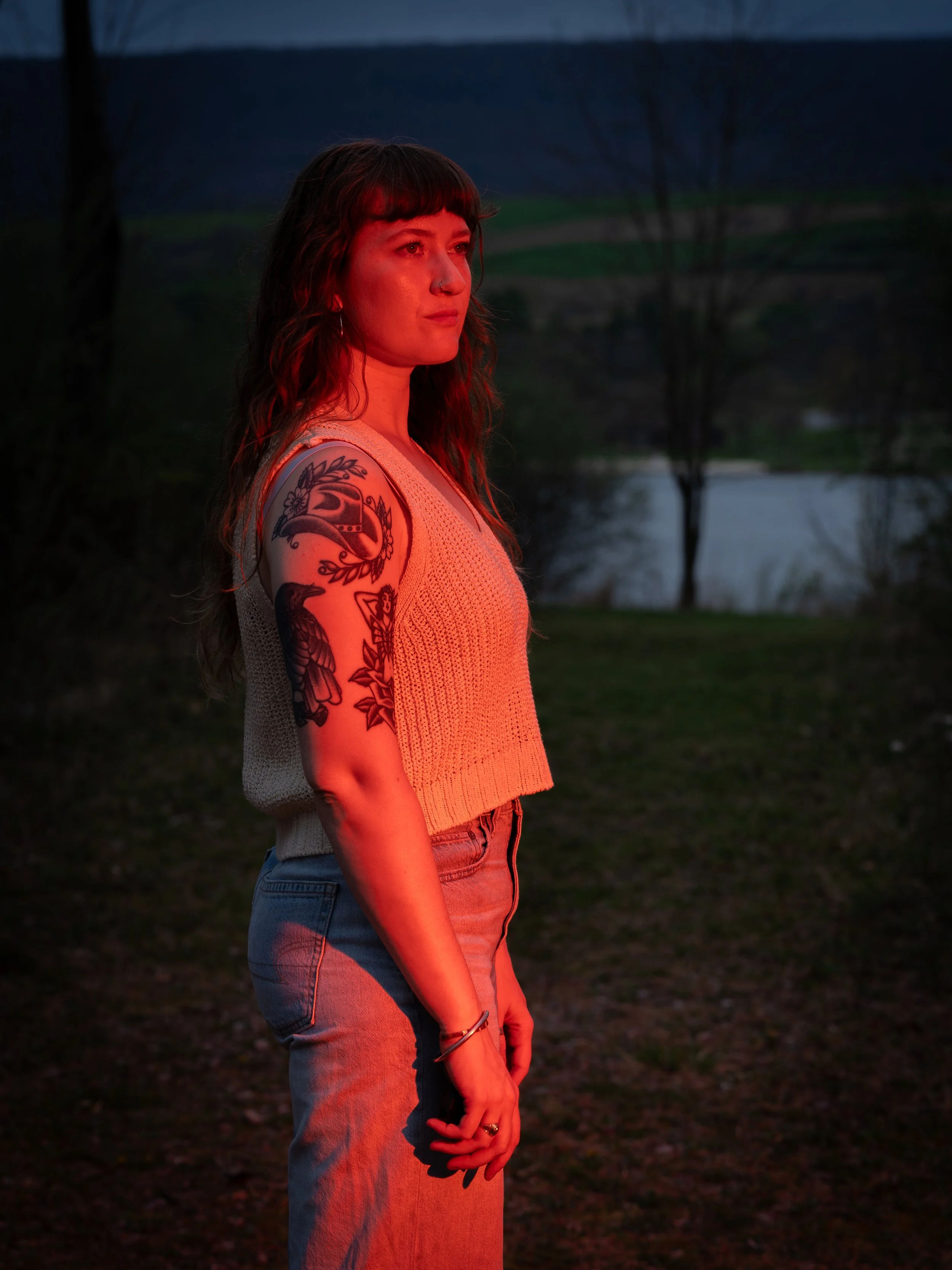 A young woman with dark hair and tattoos on her arm stands outdoors at dusk, illuminated by red light, with trees and a body of water in the background.