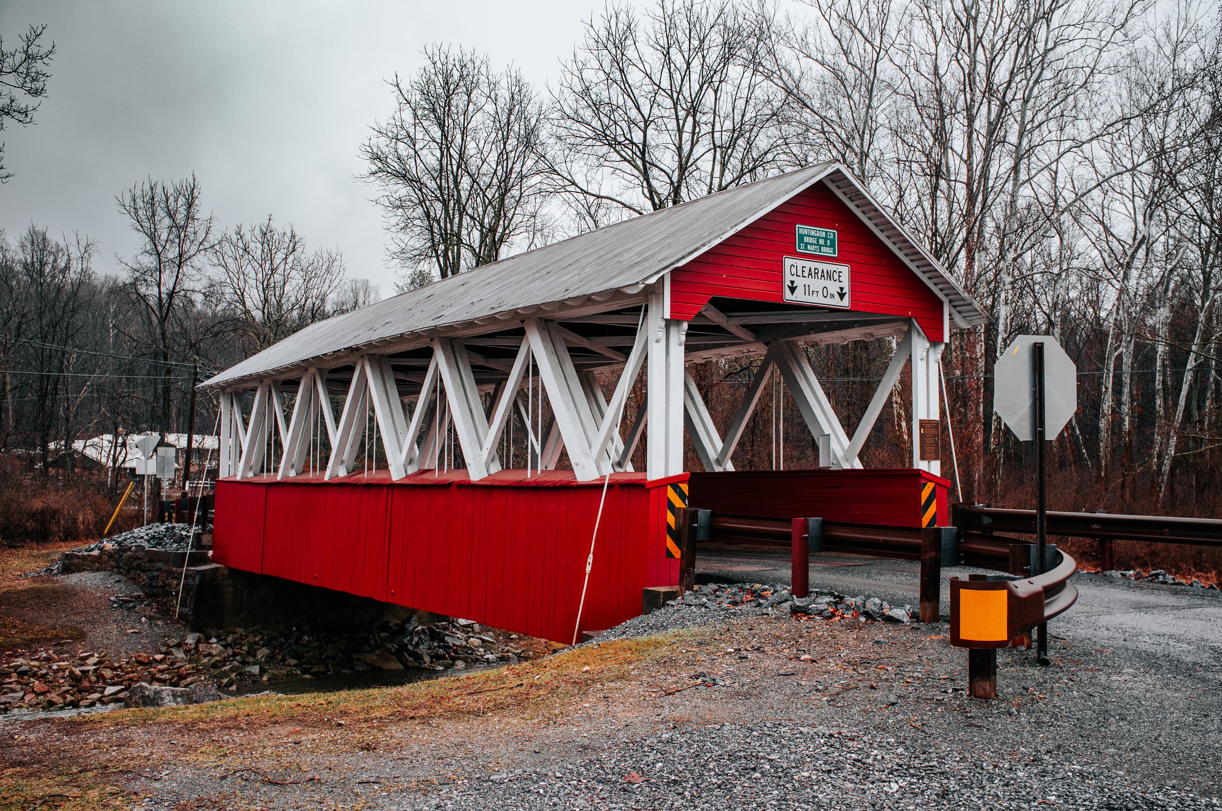 Red and white covered bridge over a small stream, with leafless trees in the background and a cloudy sky.