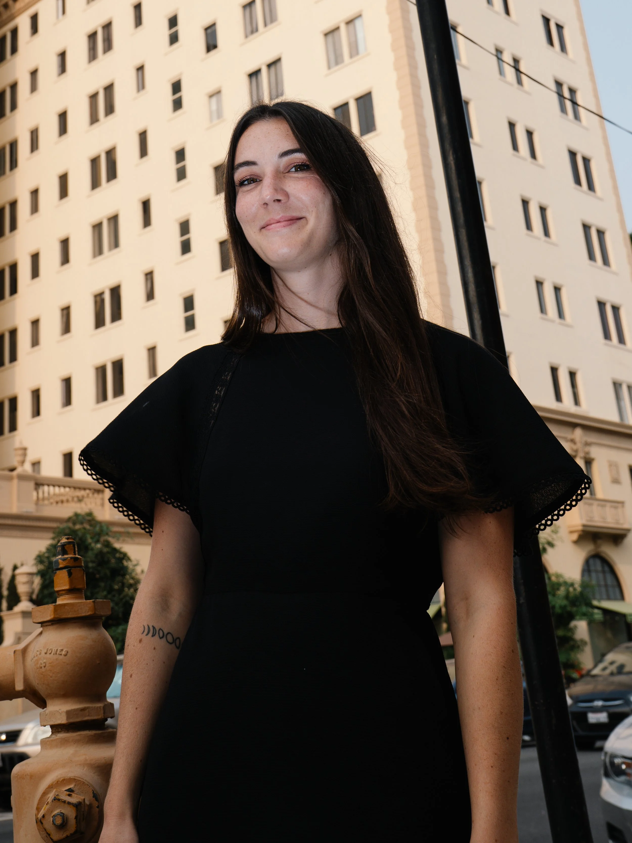 A woman with long dark hair smiling in front of a tall beige building during the day.