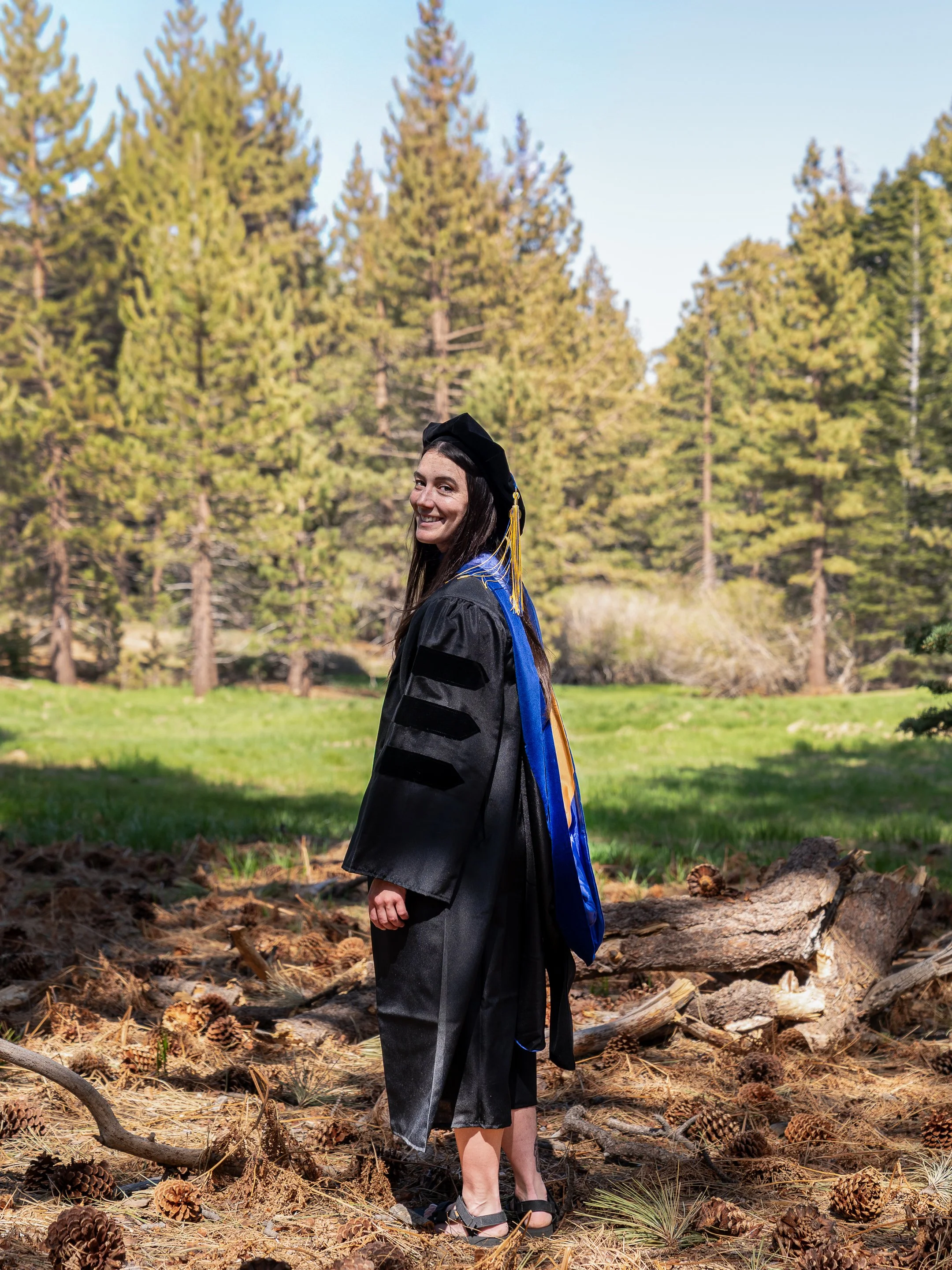 A young woman in graduation attire, including a black cap and gown, stands outdoors on a clear day among pine trees and fallen pinecones, smiling at the camera.