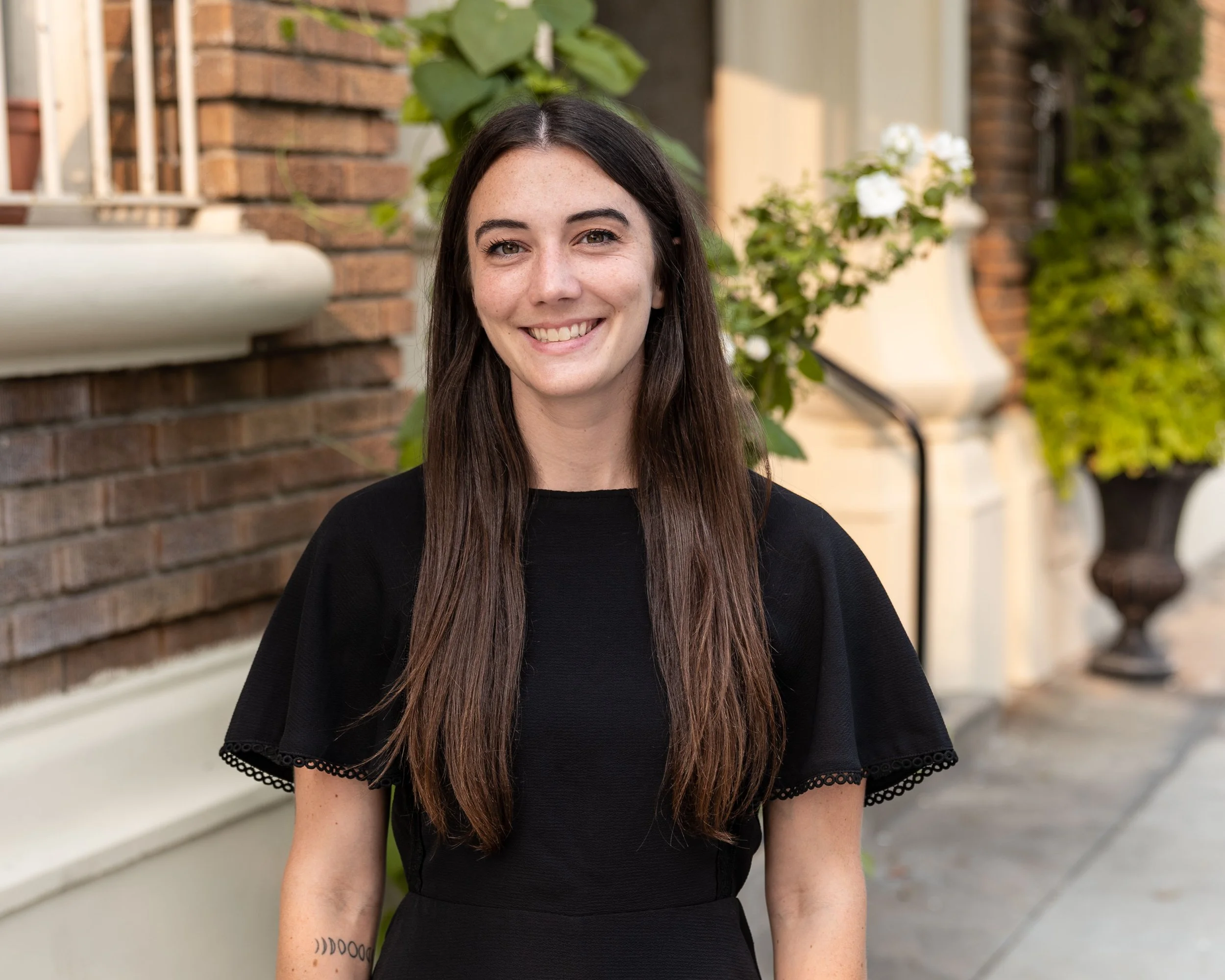 A young woman with long brown hair, wearing a black dress with lace trim, smiling outdoors in front of a brick building and greenery.