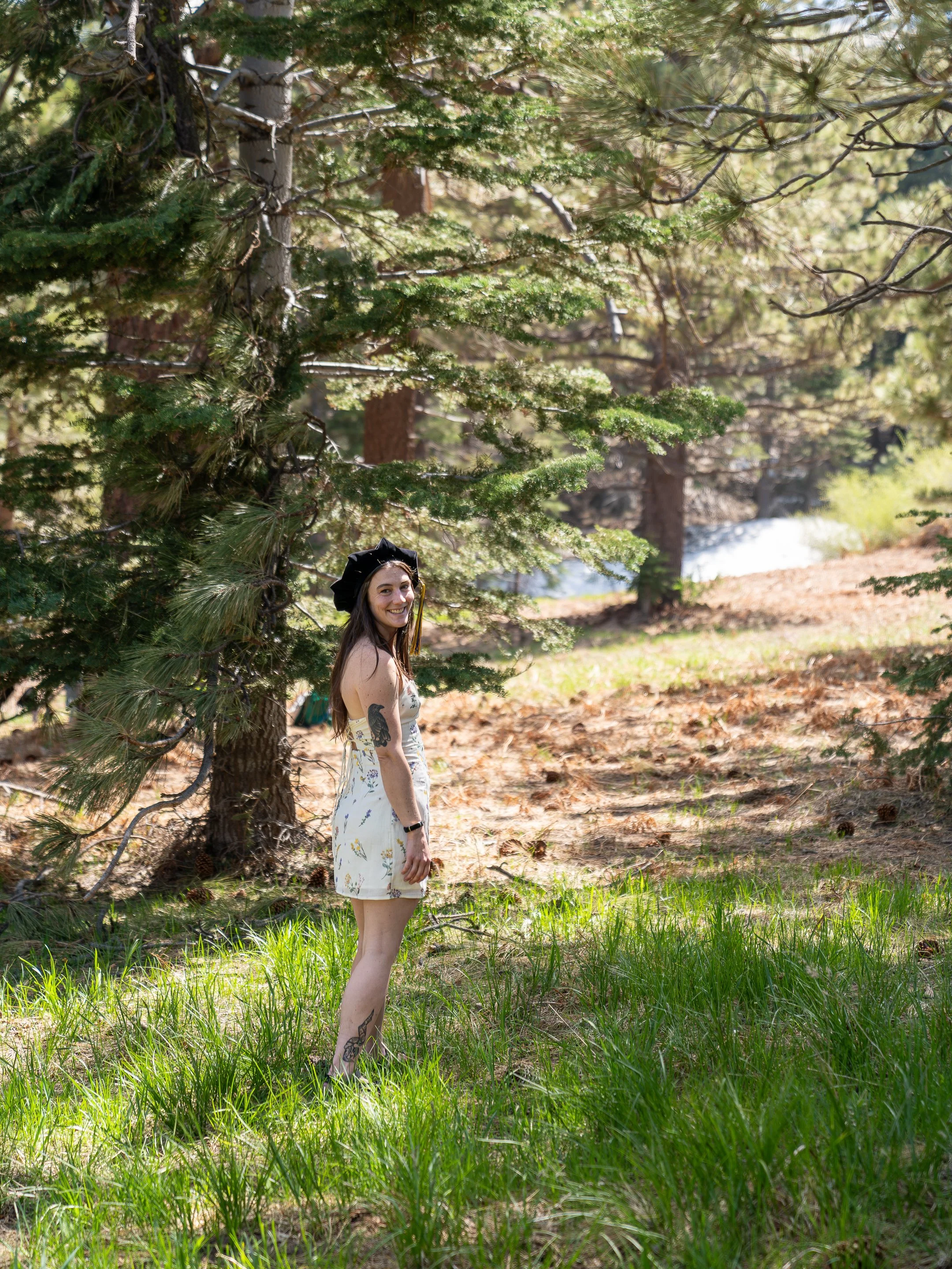A young woman with long brown hair, tattoos on her arm and leg, wearing a black cap and a white dress with yellow flowers, standing in a grassy area with a large pine tree in a forested setting during daytime.