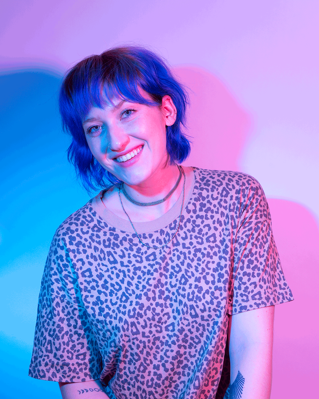 A young woman with short blue hair and a nose piercing smiling, wearing a leopard print t-shirt and layered necklaces, against a colorful pink and blue lit background.