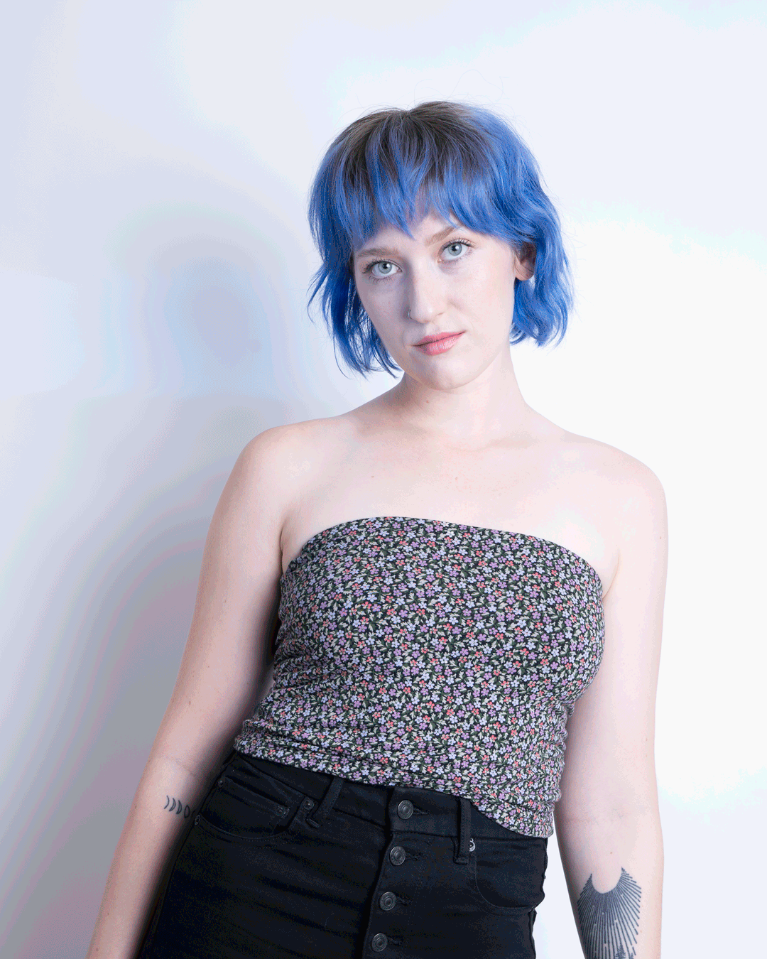 A woman with short, blue hair, wearing a strapless floral top and black pants, standing against a plain light background.