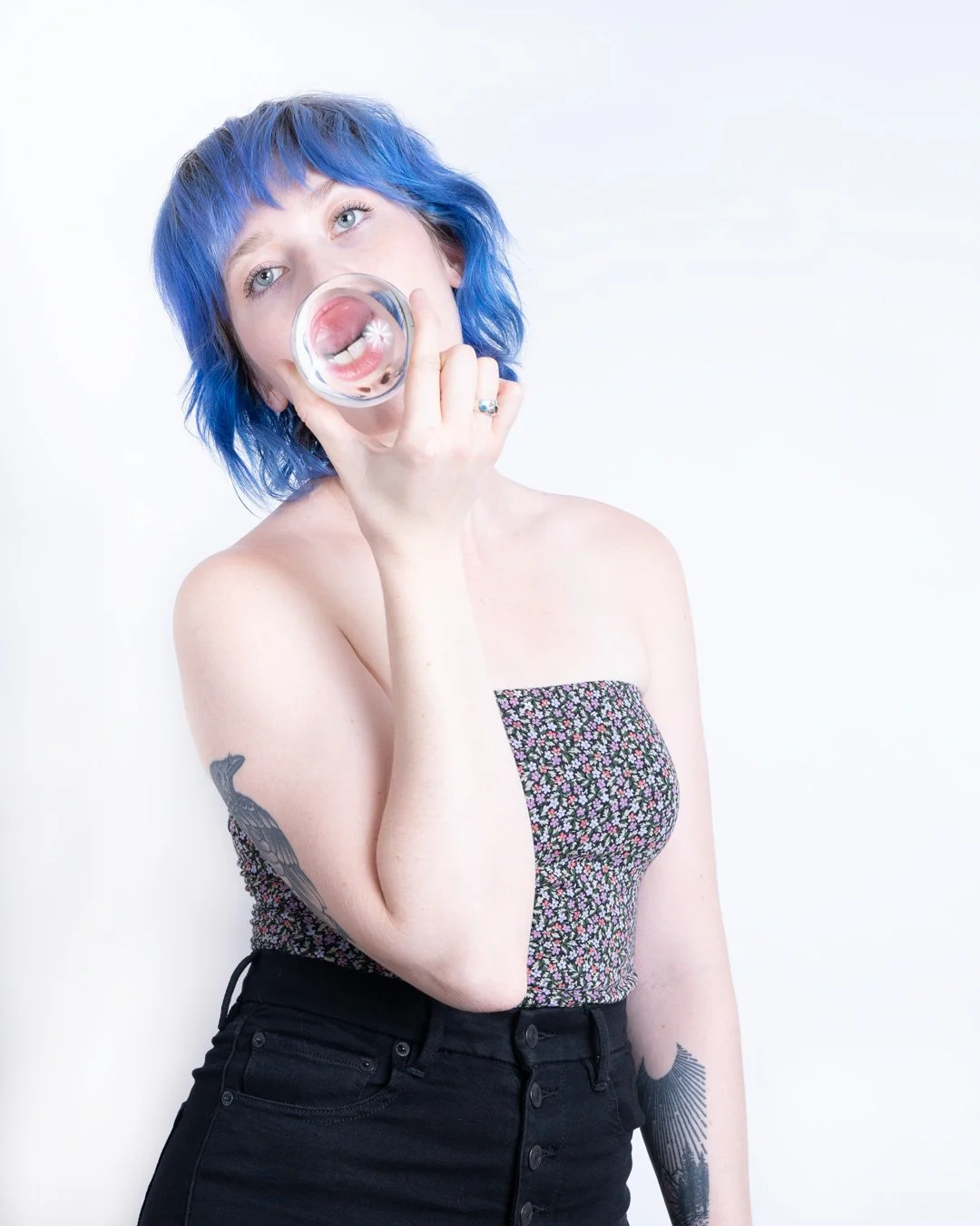 A woman with blue hair holding a glass to her mouth, creating a reflection of her face and tongue, against a plain white background.