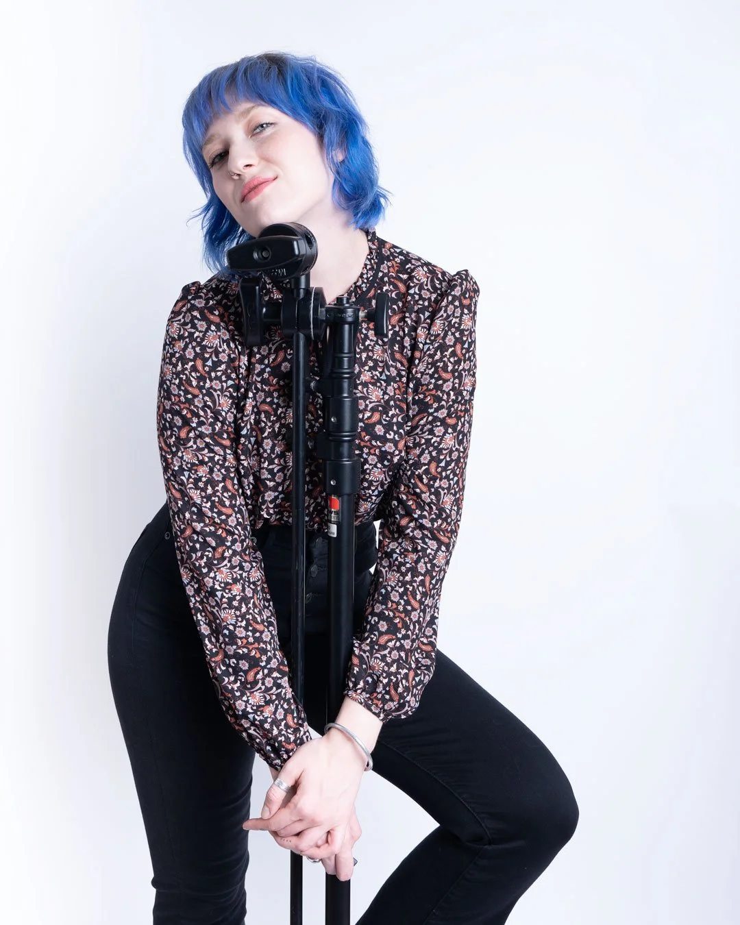 Woman with short blue hair in floral blouse posing with camera on tripod against white background.