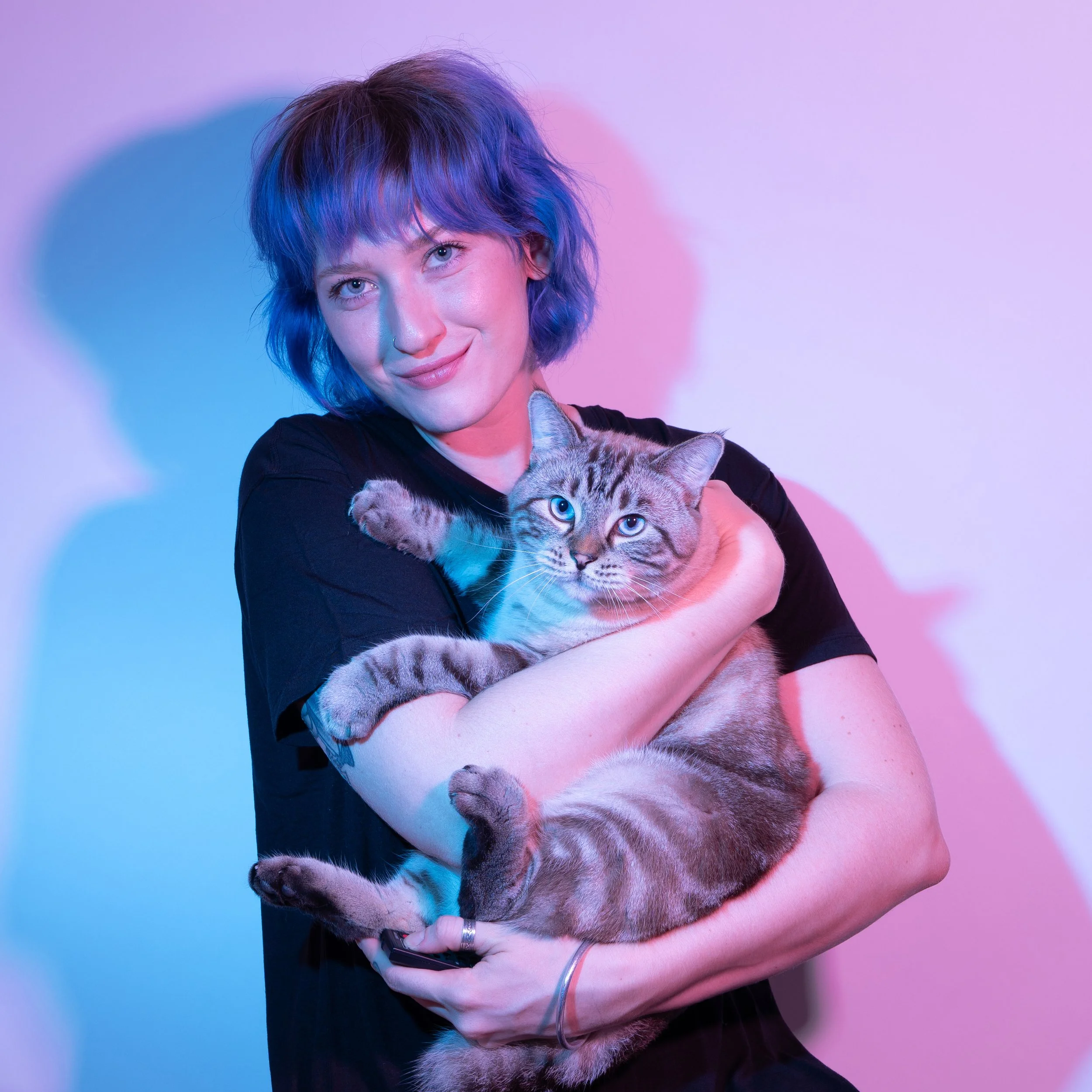 A woman with short, blue hair holding a tabby cat with light brown fur and dark stripes, against a background with pink and blue lighting.