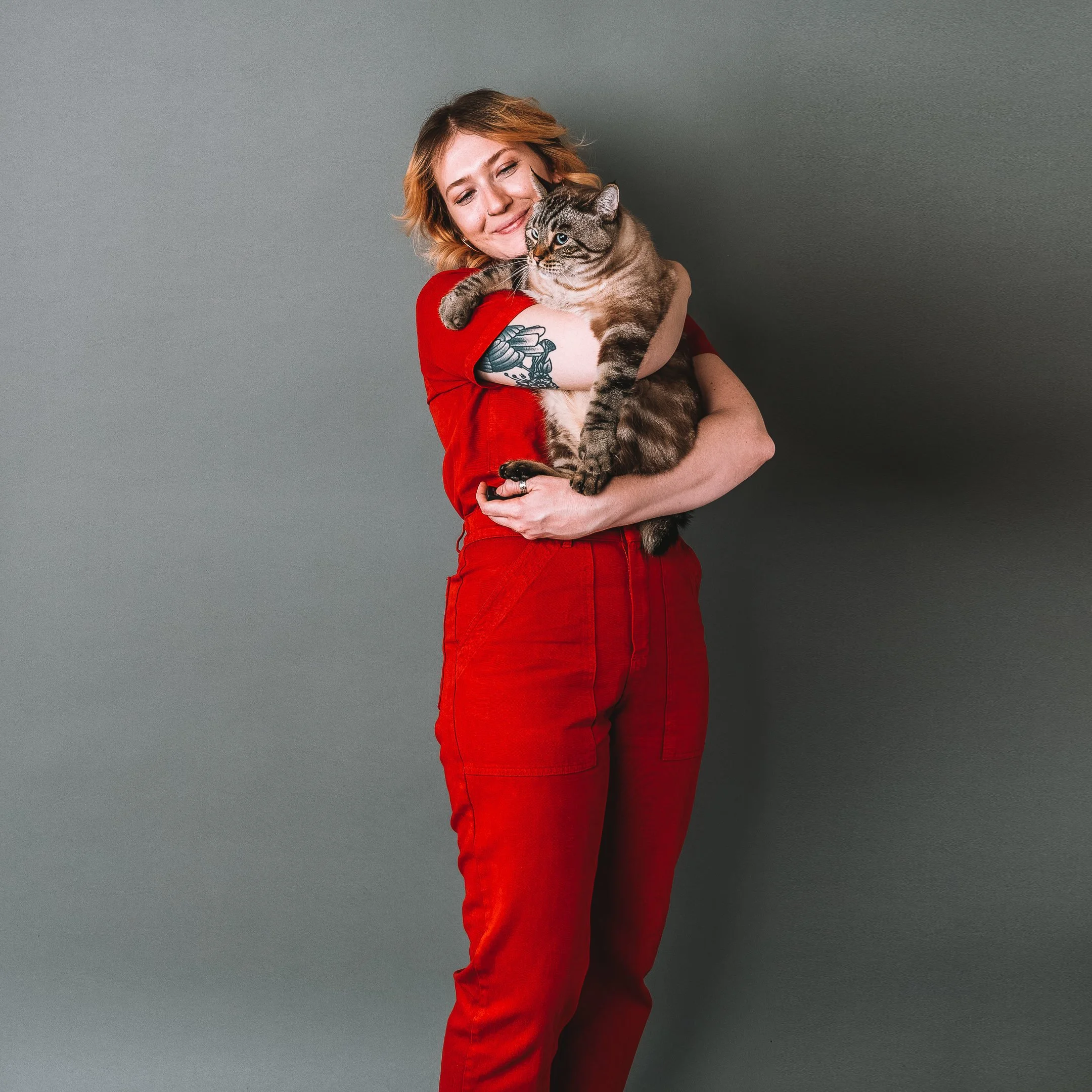 A woman with shoulder-length blonde hair wearing a red outfit, smiling with eyes closed, hugging a large tabby cat against a plain gray background.