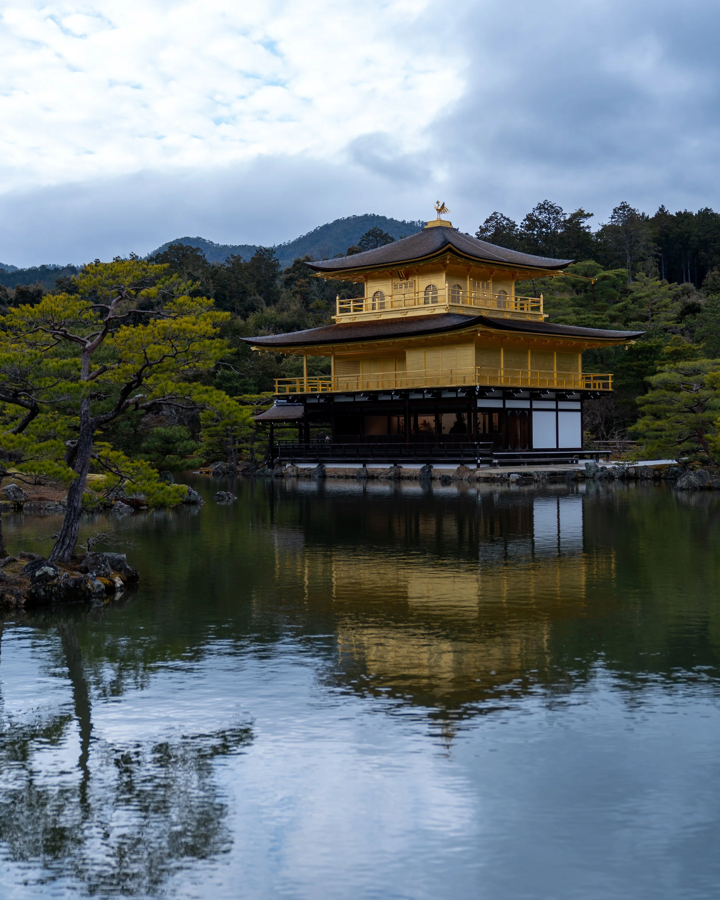 A traditional Japanese building with gold exterior and black roof, reflecting in a pond surrounded by trees and mountains under a cloudy sky.