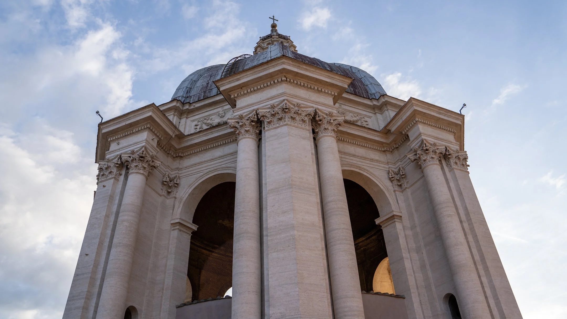 View of a historic church or cathedral with tall columns, detailed architecture, and a dome topped with a cross, set against a partly cloudy sky.