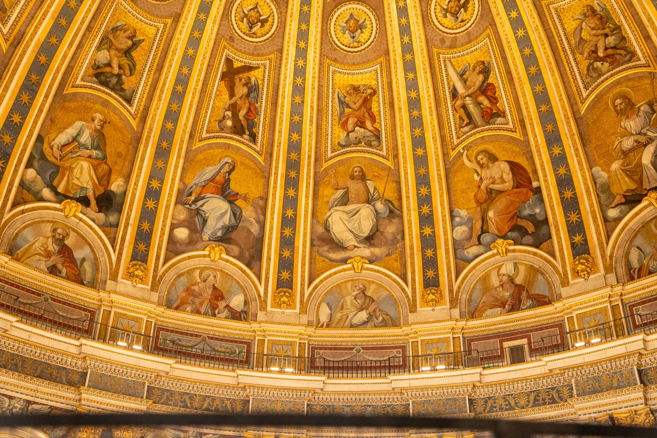 The ornate ceiling of a church, decorated with golden embellishments and religious paintings depicting various biblical figures and scenes, arranged in a circular pattern.