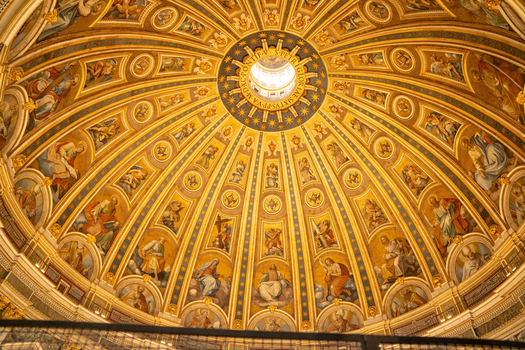 A grand, ornate domed ceiling with gold accents and religious artwork depicting various saints and biblical figures, with a Latin inscription around the central opening.
