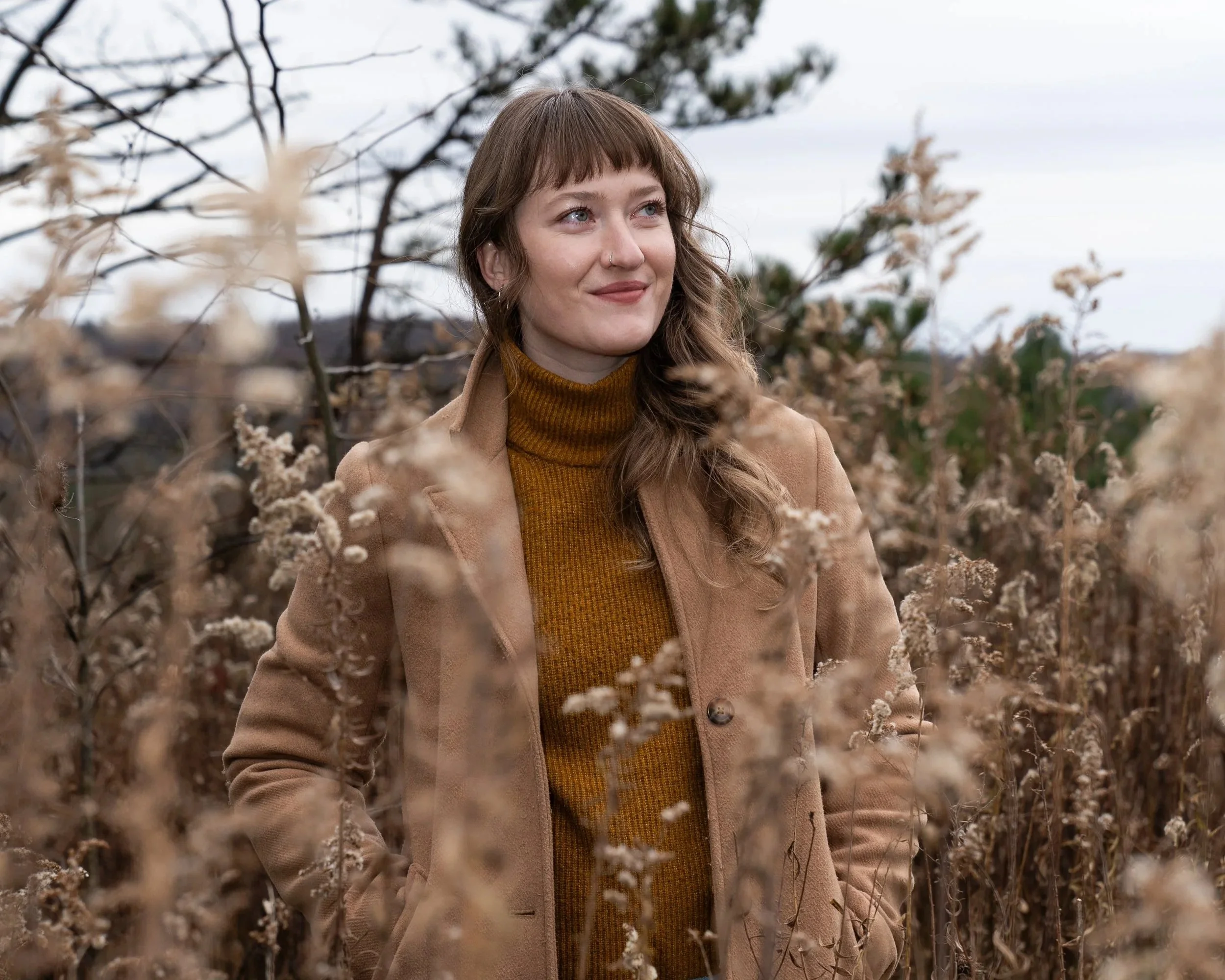 A woman with light skin, brown hair, and a nose piercing standing among dry plants outdoors on a cloudy day.